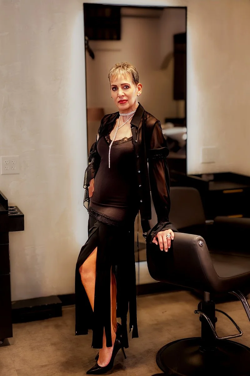 A woman with short blonde hair wearing black lace top, sheer black skirt with high slit, black heels, and jewelry, standing in a room with a mirror, chair, and desk.