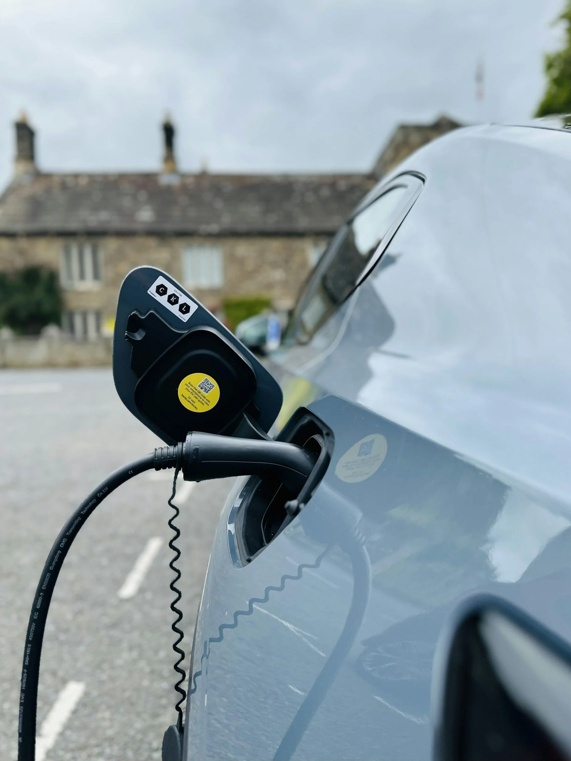 Electric vehicle charging port with charging cable plugged in, set against a blurry background of a stone house and cloudy sky.