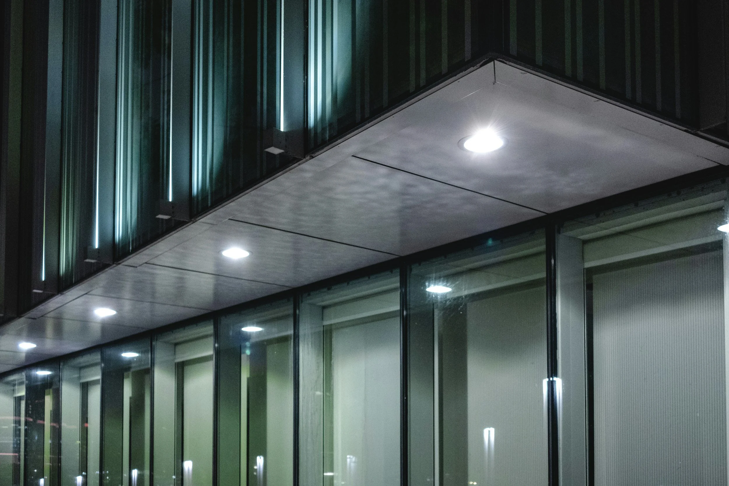 Nighttime view of a modern building's exterior with glass windows and illuminated ceiling lights reflecting in the glass.