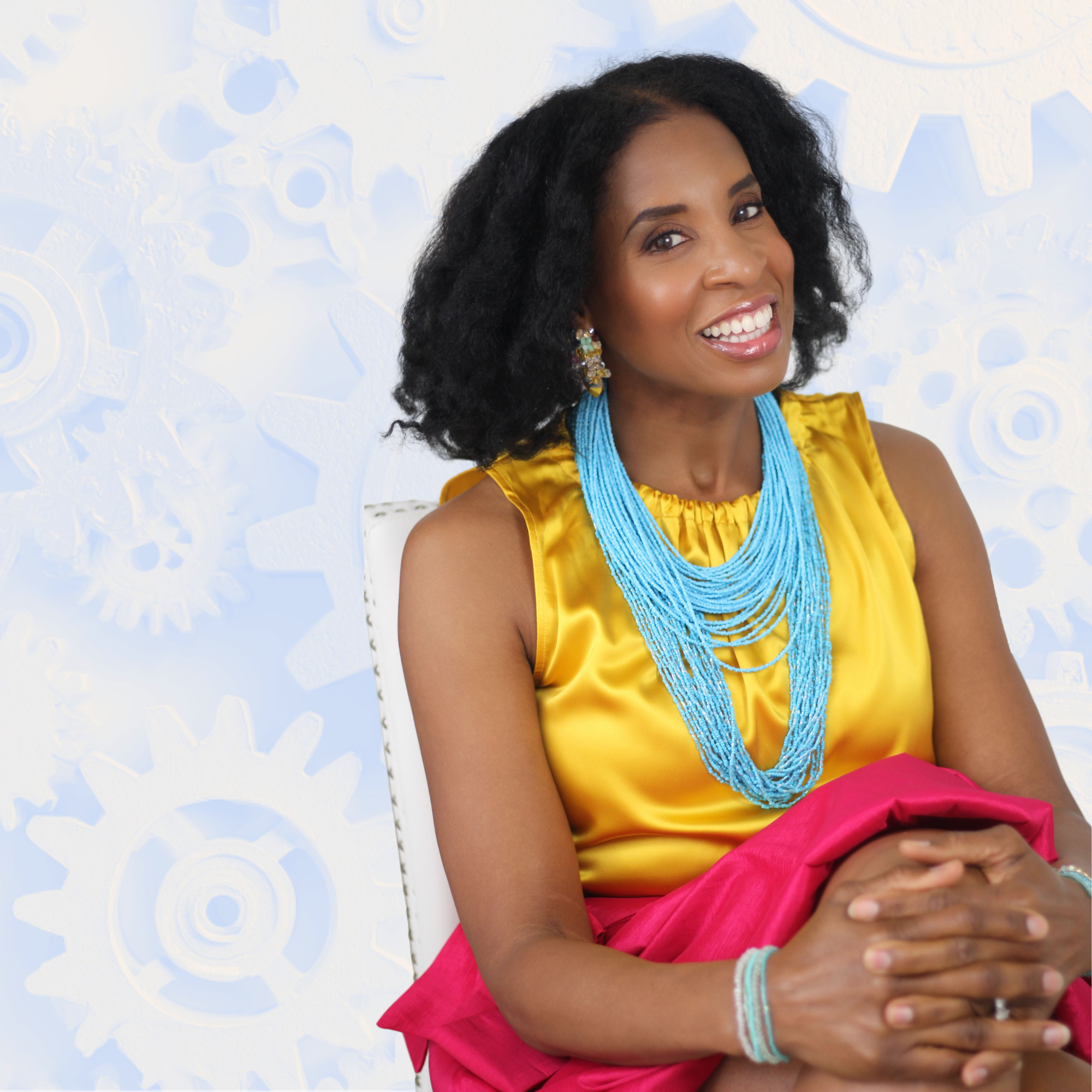 Dara Fairman wearing a yellow satin top, a pink skirt, and layered turquoise necklaces, sitting with her hands clasped and smiling. She has black curly hair and wears earrings and bracelets, with a background of white gears and cogs