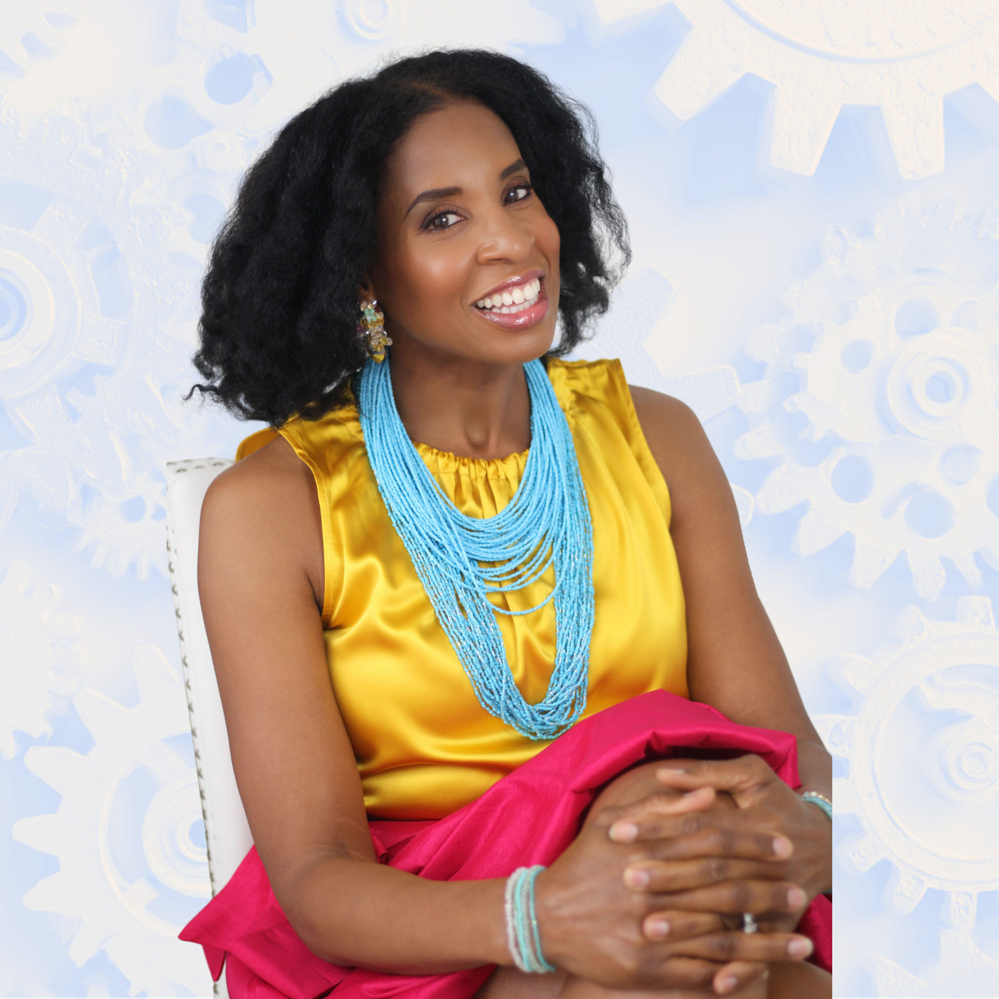 Dara Fairman, smiling, wearing a yellow satin top and a bright pink skirt, accessorized with multiple blue beaded necklaces, floral earrings, and a bracelet, sitting with her hands clasped.