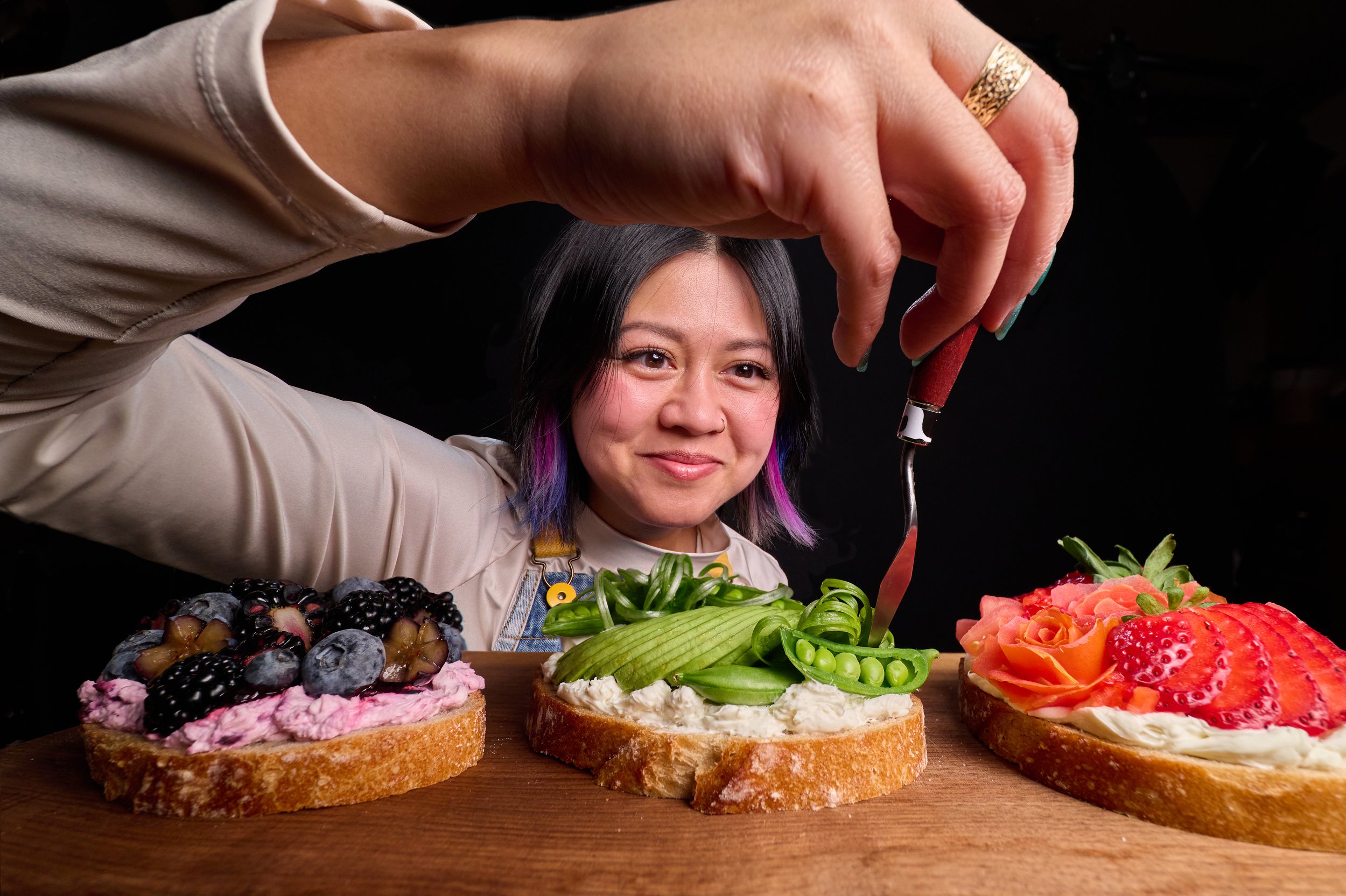 A woman smiling as she decorates fruit-topped cakes with a piping tool on a wooden surface, against a dark background.