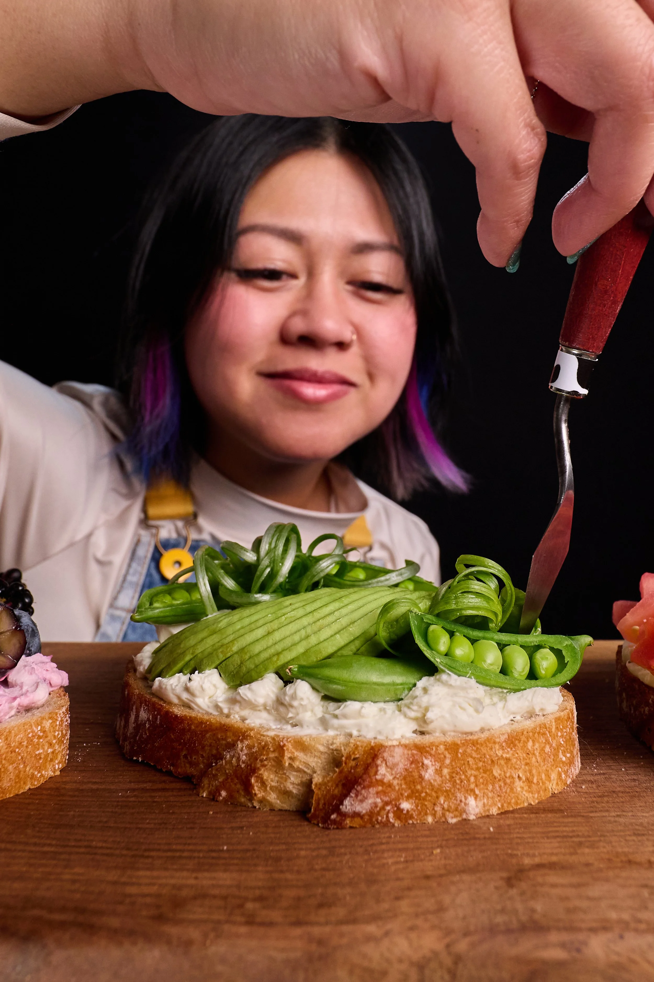 A woman with black hair and purple tips decorating a slice of bread with cream cheese and green vegetables, with a smile and a focused expression.
