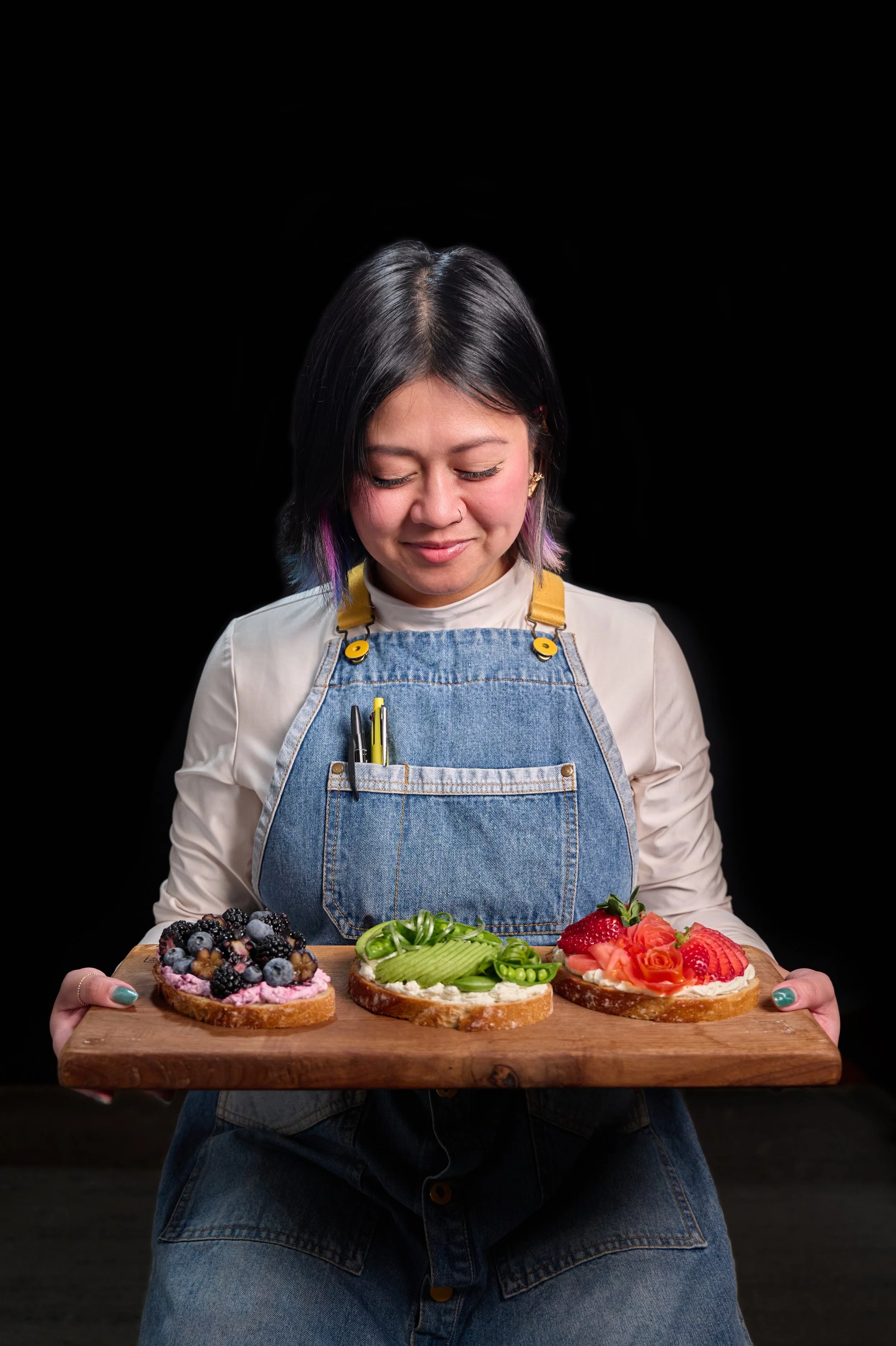 A woman holding a wooden platter with three fruit-topped open-faced sandwiches, smiling gently and looking down at the food, against a black background.