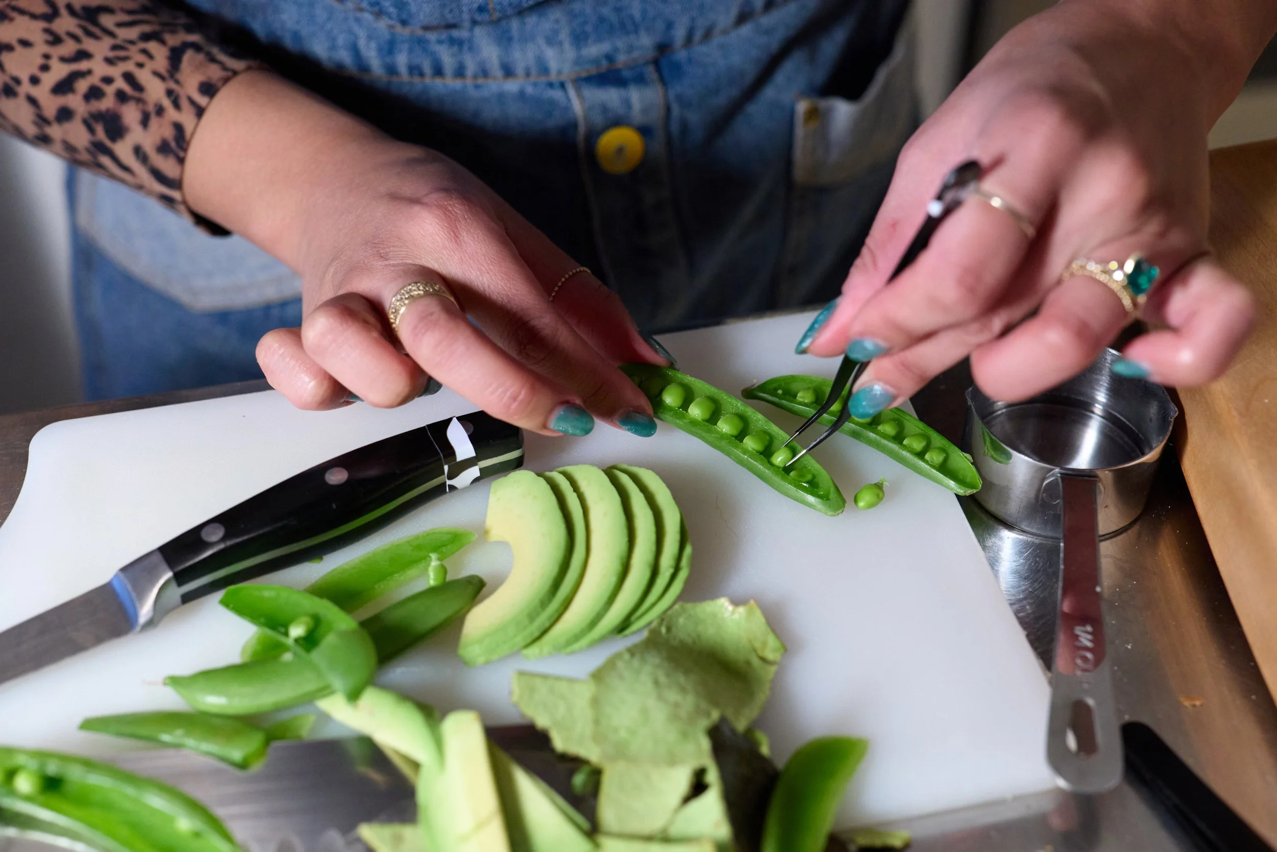 A person with multiple rings on their fingers is using tweezers to remove seeds from an open pea pod on a white cutting board. There are sliced avocado and other green vegetables on the cutting board, with a knife nearby.