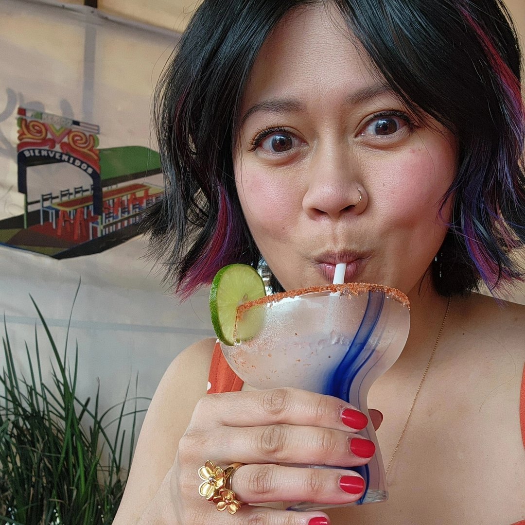 Woman with black and purple hair drinking a margarita with a lime wedge, salt rim, and blue stirrer.