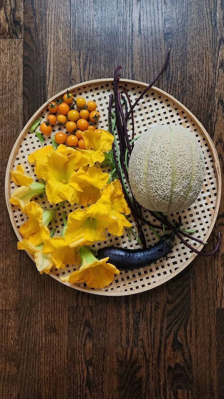 A round woven basket with a lattice pattern contains various vegetables including yellow squash, small orange cherry tomatoes, long purple eggplants, a large white cantaloupe, and a few green beans, placed on a dark wood surface.