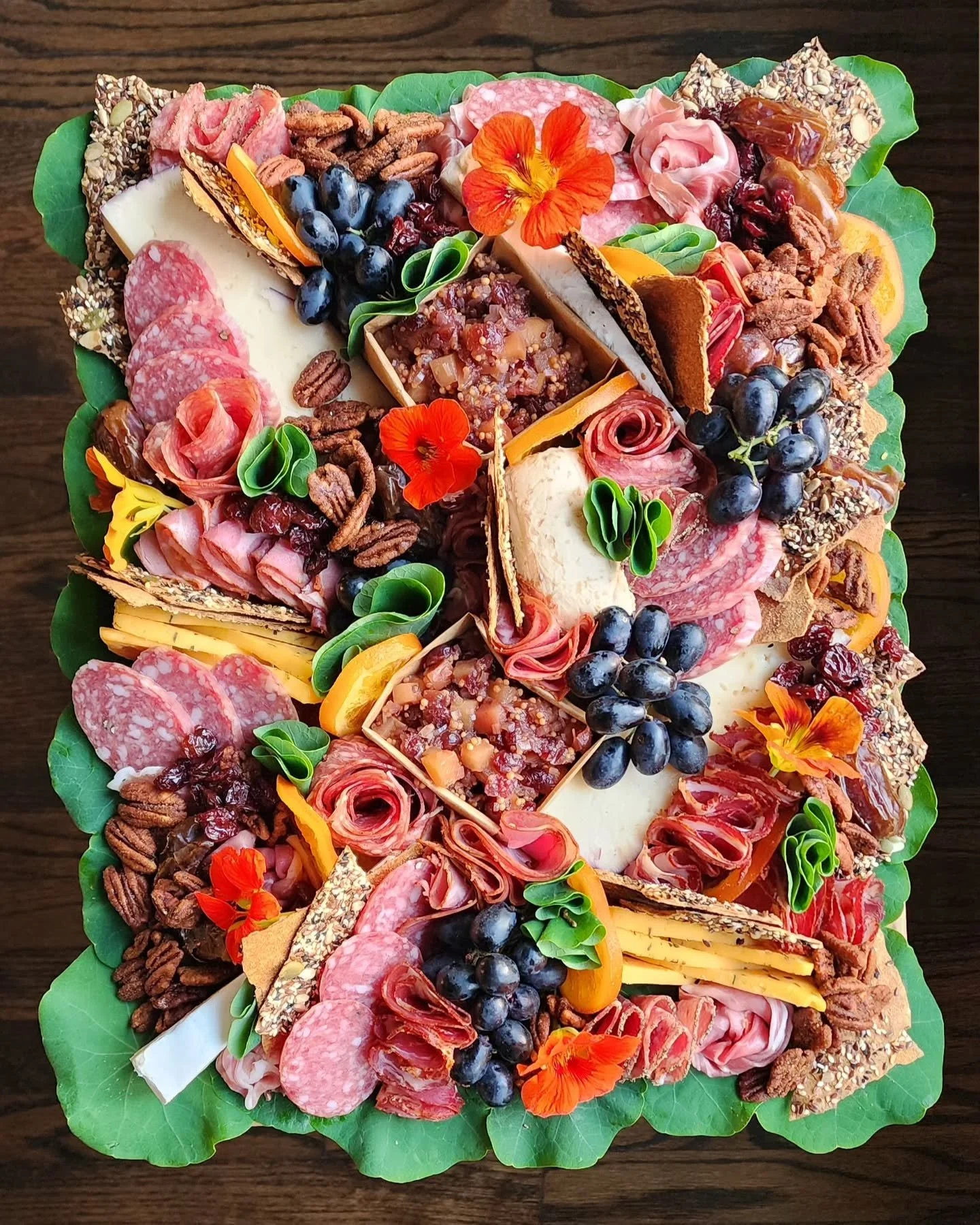 A large charcuterie board with assorted cheeses, cured meats, grapes, nuts, crackers, and edible flowers on a wooden surface.