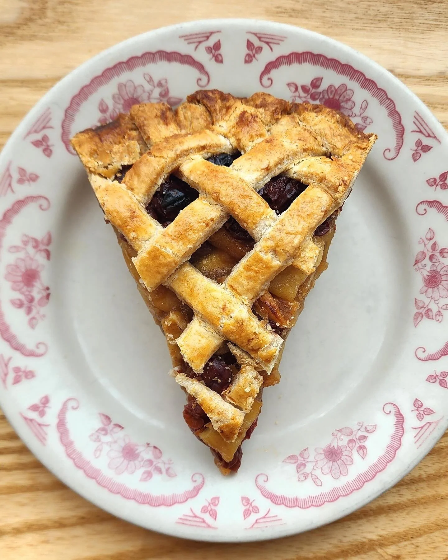 Slice of berry pie with lattice crust on a decorative white plate with red floral designs, placed on a wooden surface.