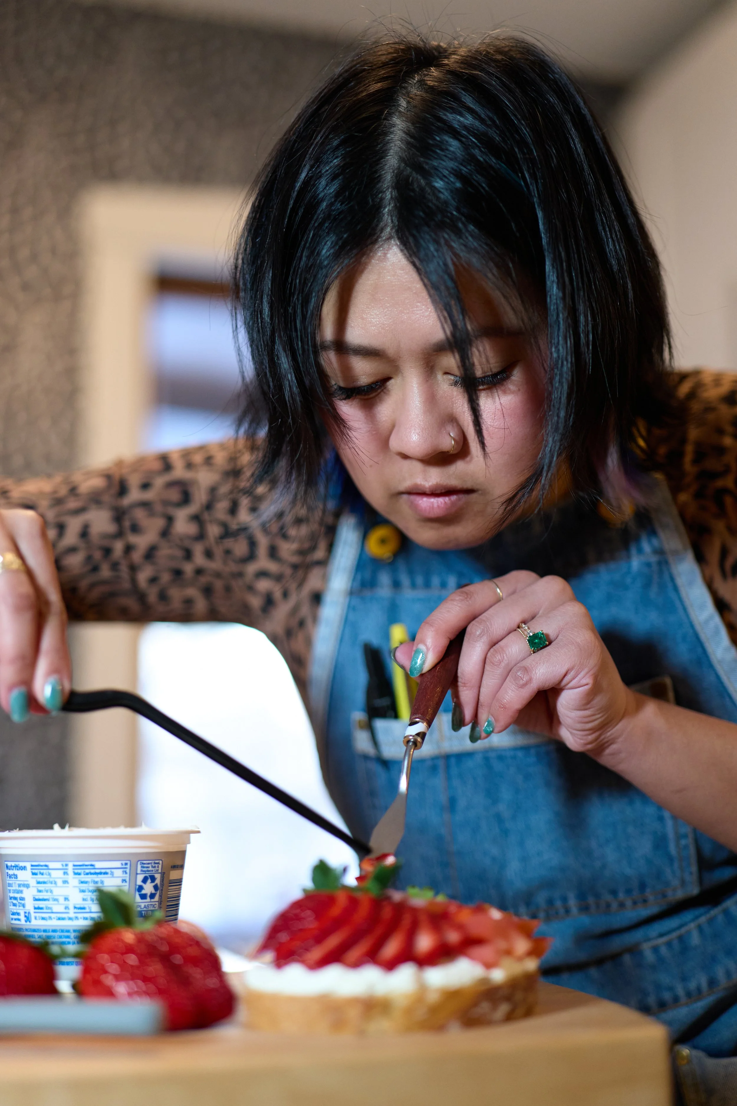 A woman with black hair and a nose ring, wearing a leopard print top and denim apron, decorating a strawberry tart with fruit and cream on a wooden surface.