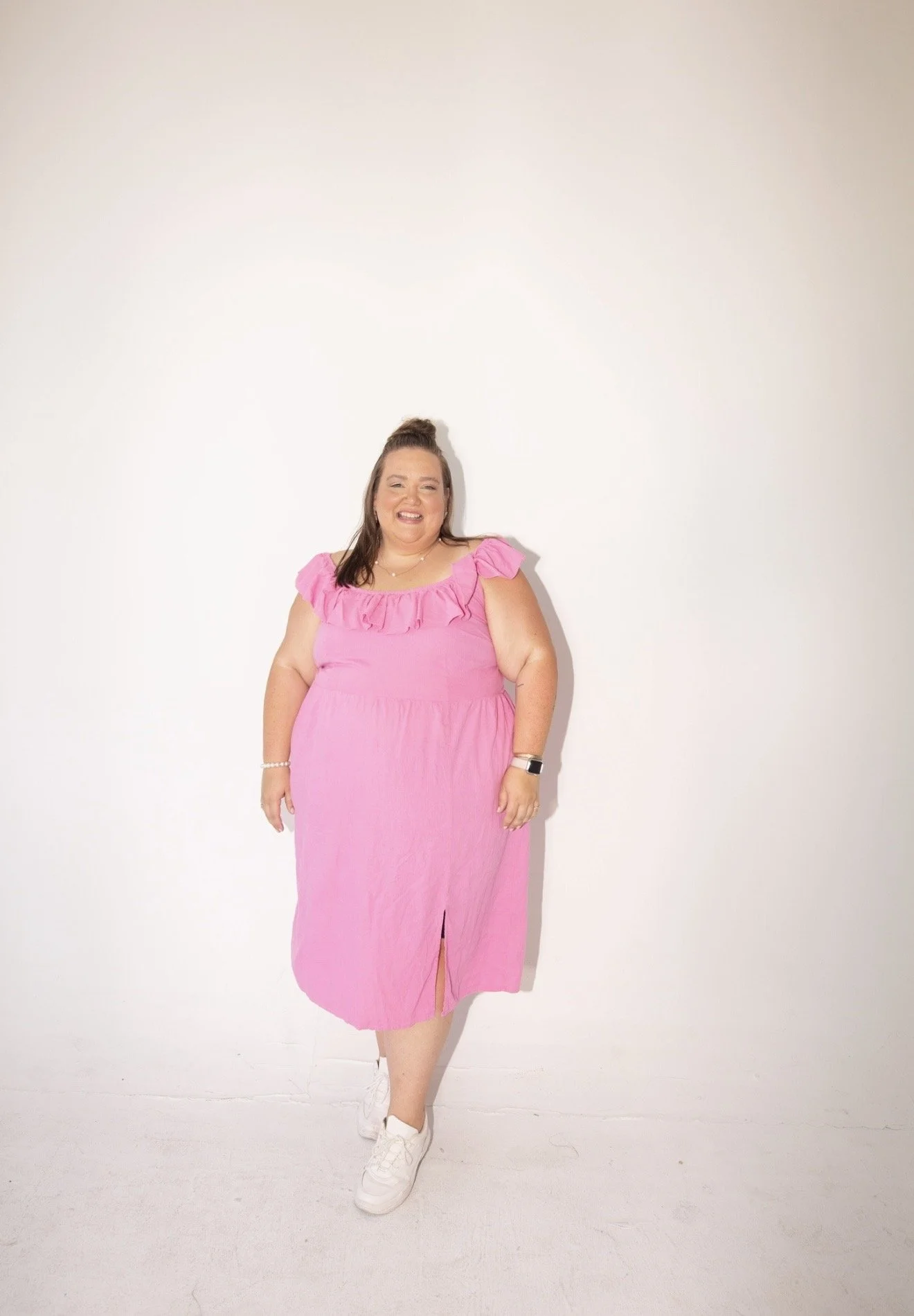 A woman standing against a plain white wall, wearing a pink dress and white sneakers, smiling and posing for the photo.