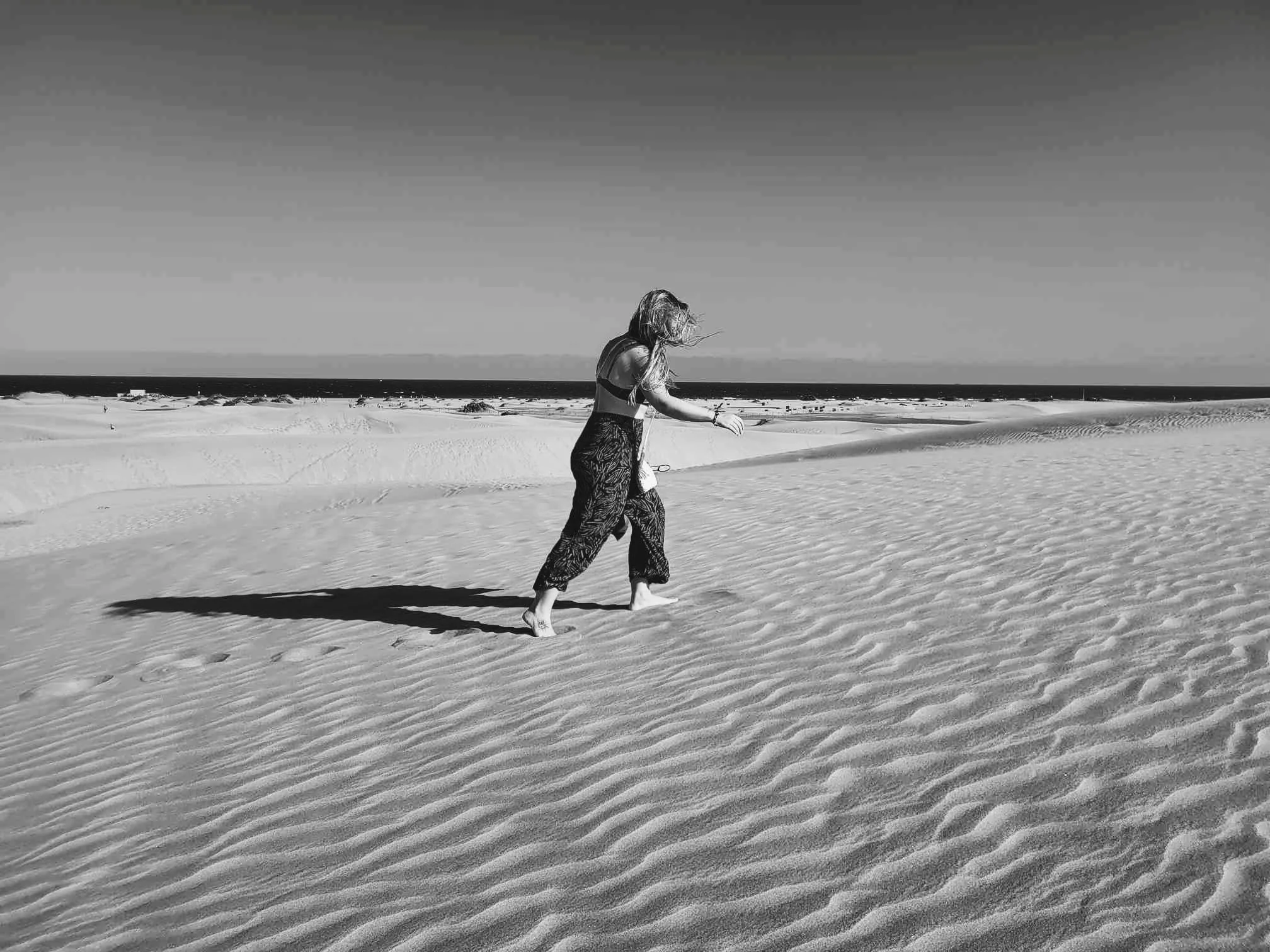 Danaé Bibeau marche pieds nus sur une plage de sable avec des dunes, sous un ciel clair.