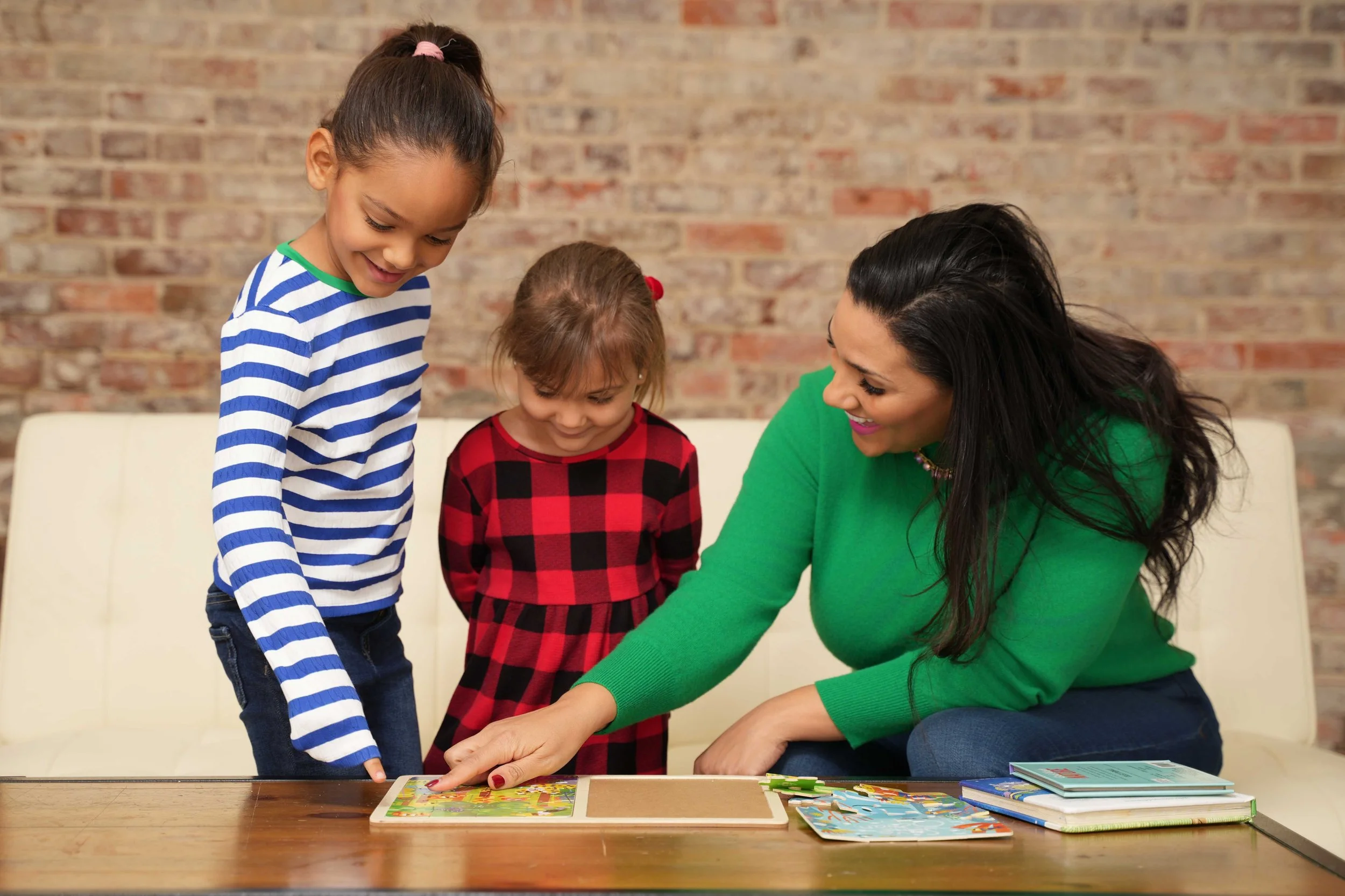 A woman with long black hair and a green sweater sitting at a table with two young girls, one in a blue and white striped shirt and the other in a red and black checkered dress, playing a board game together in a room with a brick wall background.