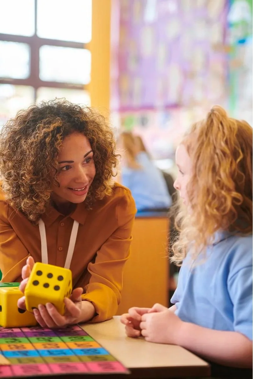 A contract speech therapist with curly hair and a mustard-colored jacket playing a board game to work on speech skills with a young girl sitting at a classroom table, providing school speech therapy services in Laurinburg, NC.