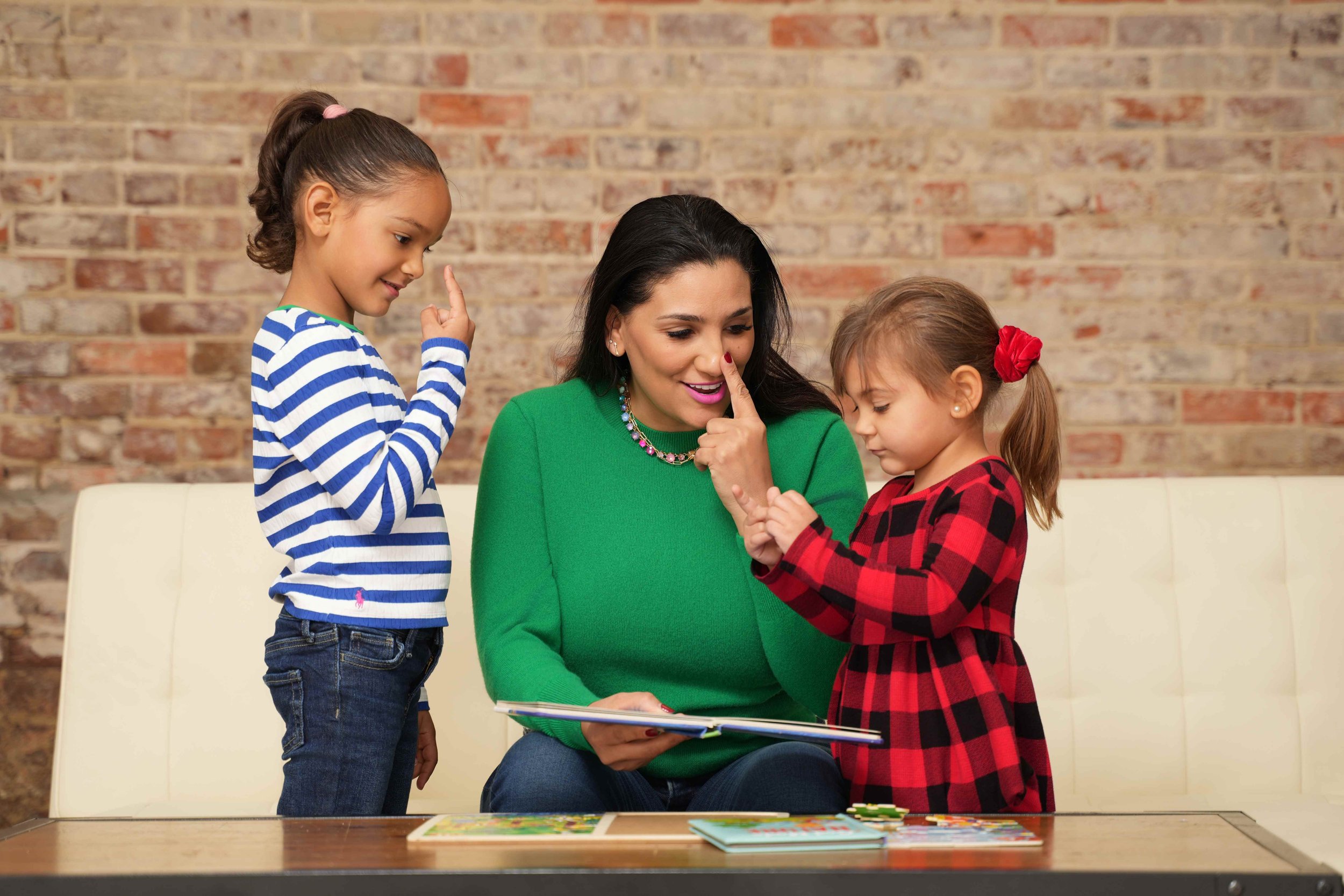 A speech therapist in a green sweater is sitting at a table with two young girls, engaging in a storytelling activity with a book during speech therapy for children in Laurinburg, NC.