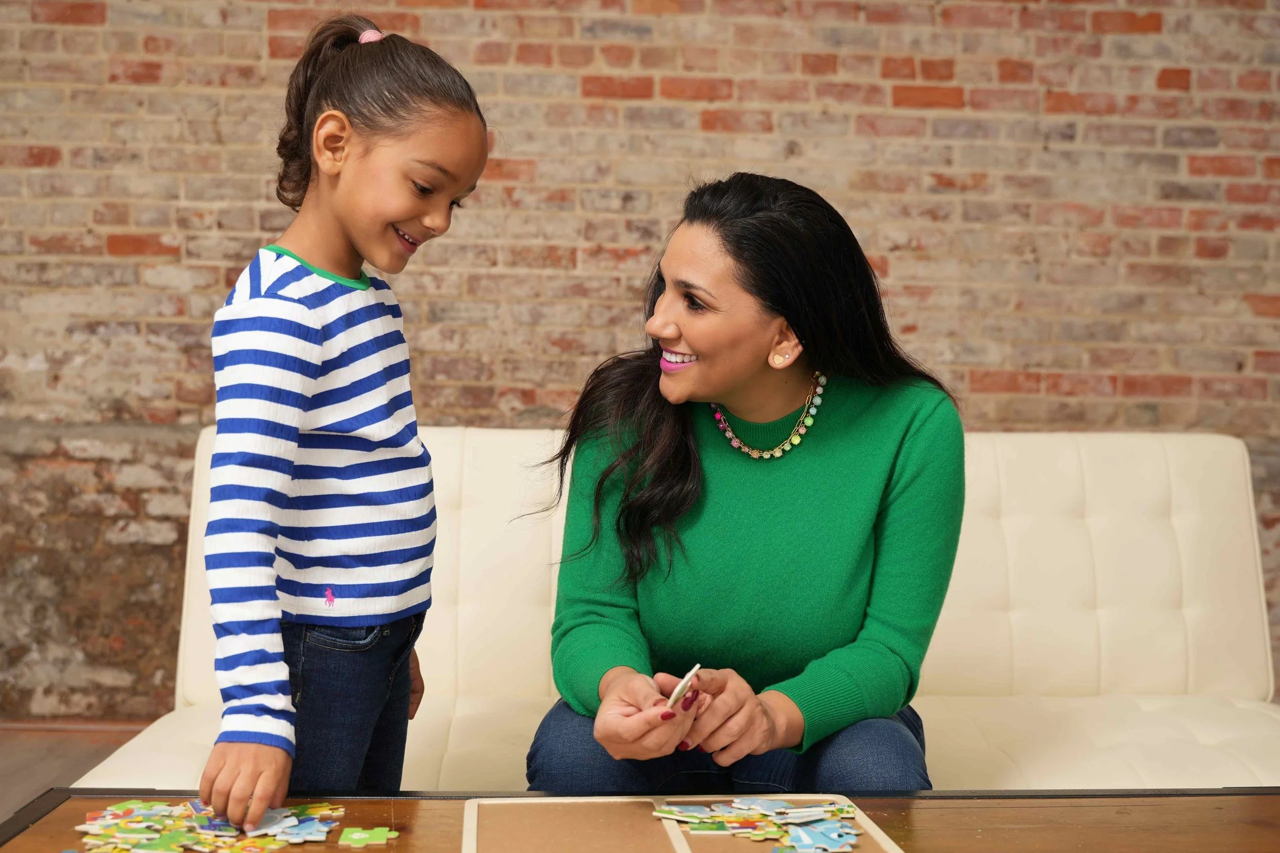A speech therapist and a young girl are sitting on a cream sofa with a brick wall in the background while using a puzzle to work on turn-taking and social skills during speech therapy for kids in Laurinburg, NC.