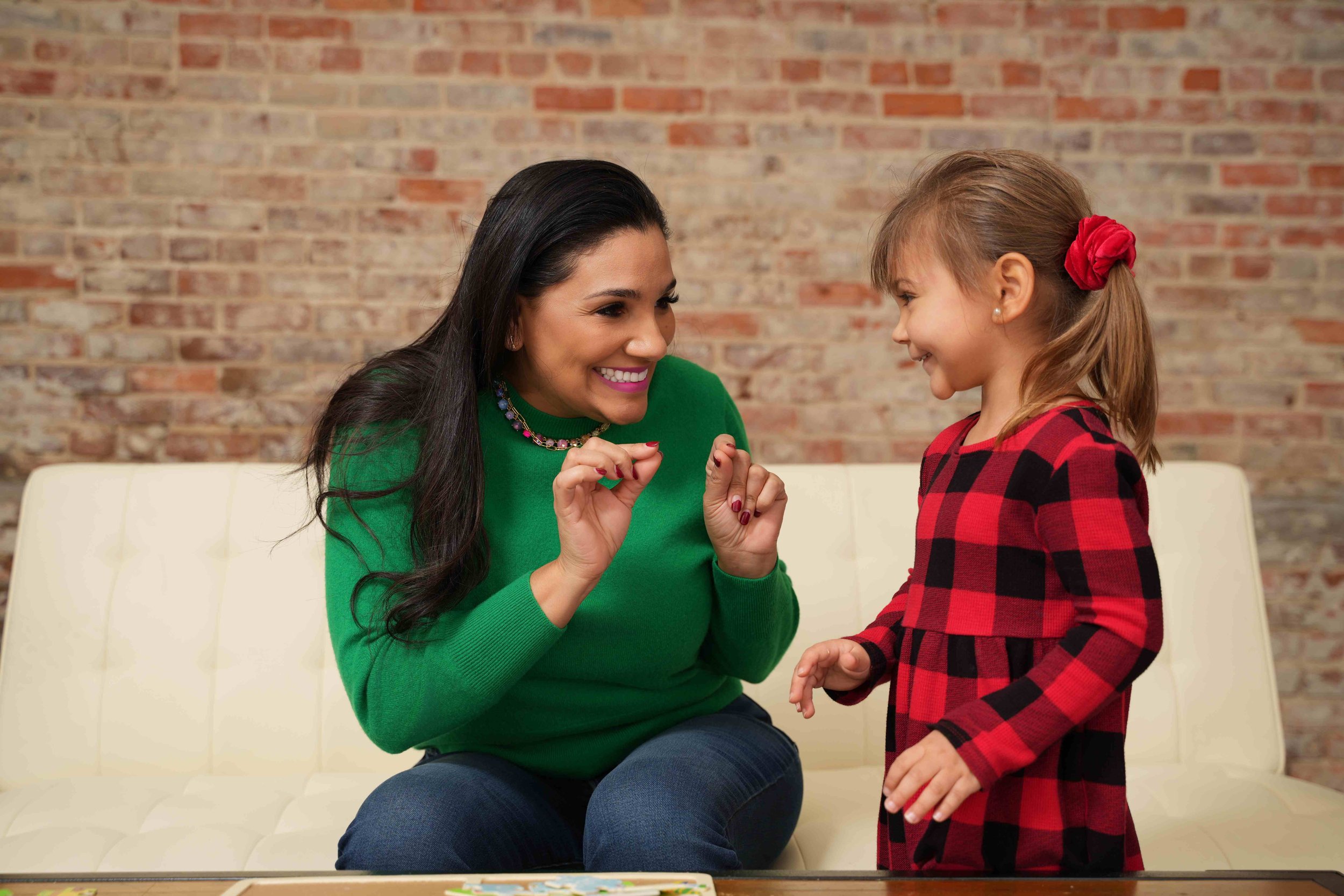 A woman and a young girl sitting on a white couch in front of a brick wall, smiling while working on speech sounds during speech therapy for kids in Laurinburg, NC.