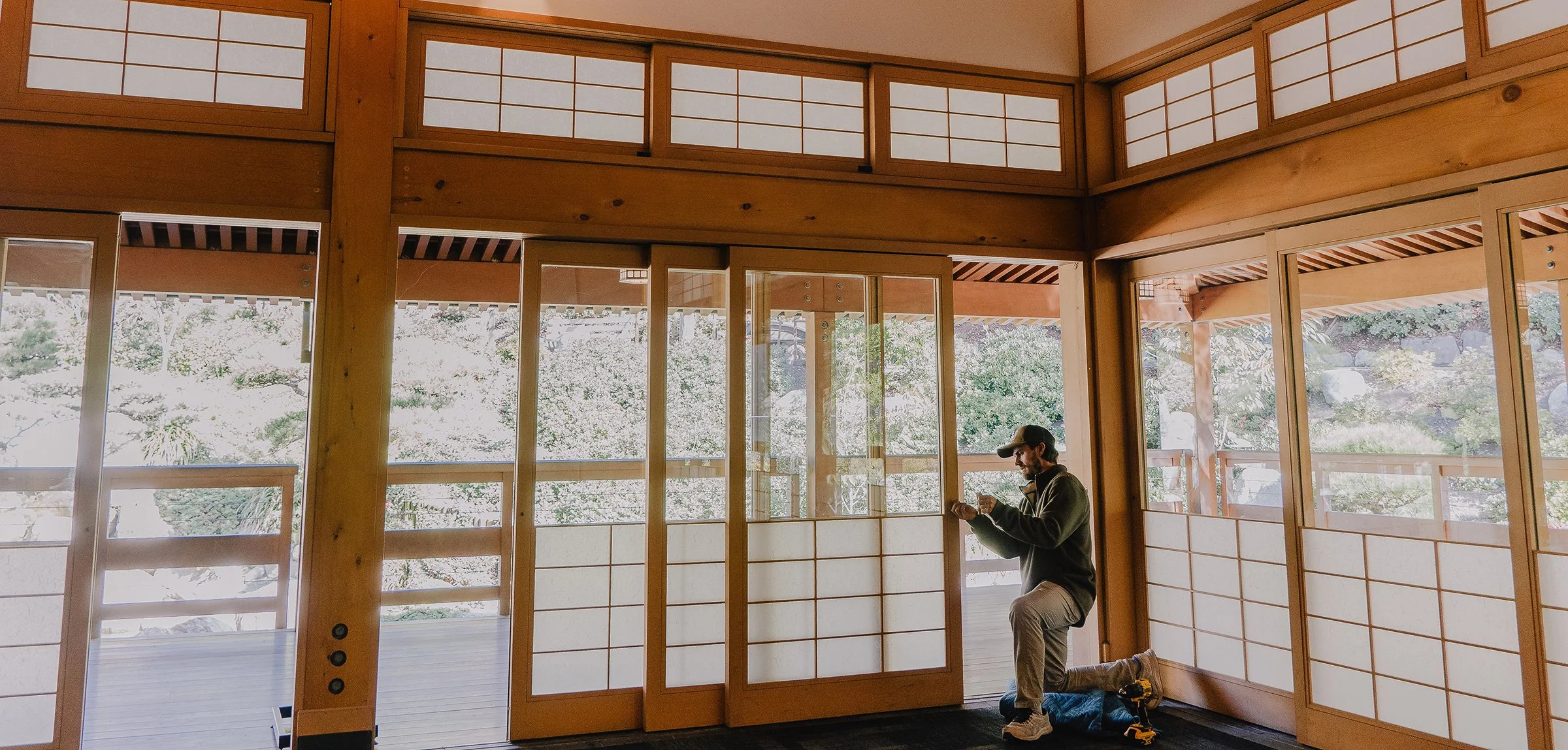 A man kneeling on the floor in a room with large windows and traditional Japanese sliding doors, working on a construction or renovation project.