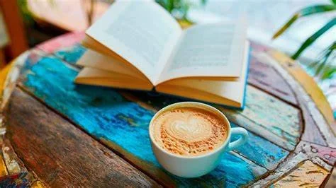 An open book and a cup of coffee on a colorful wooden table with a blurred background and green leaves.