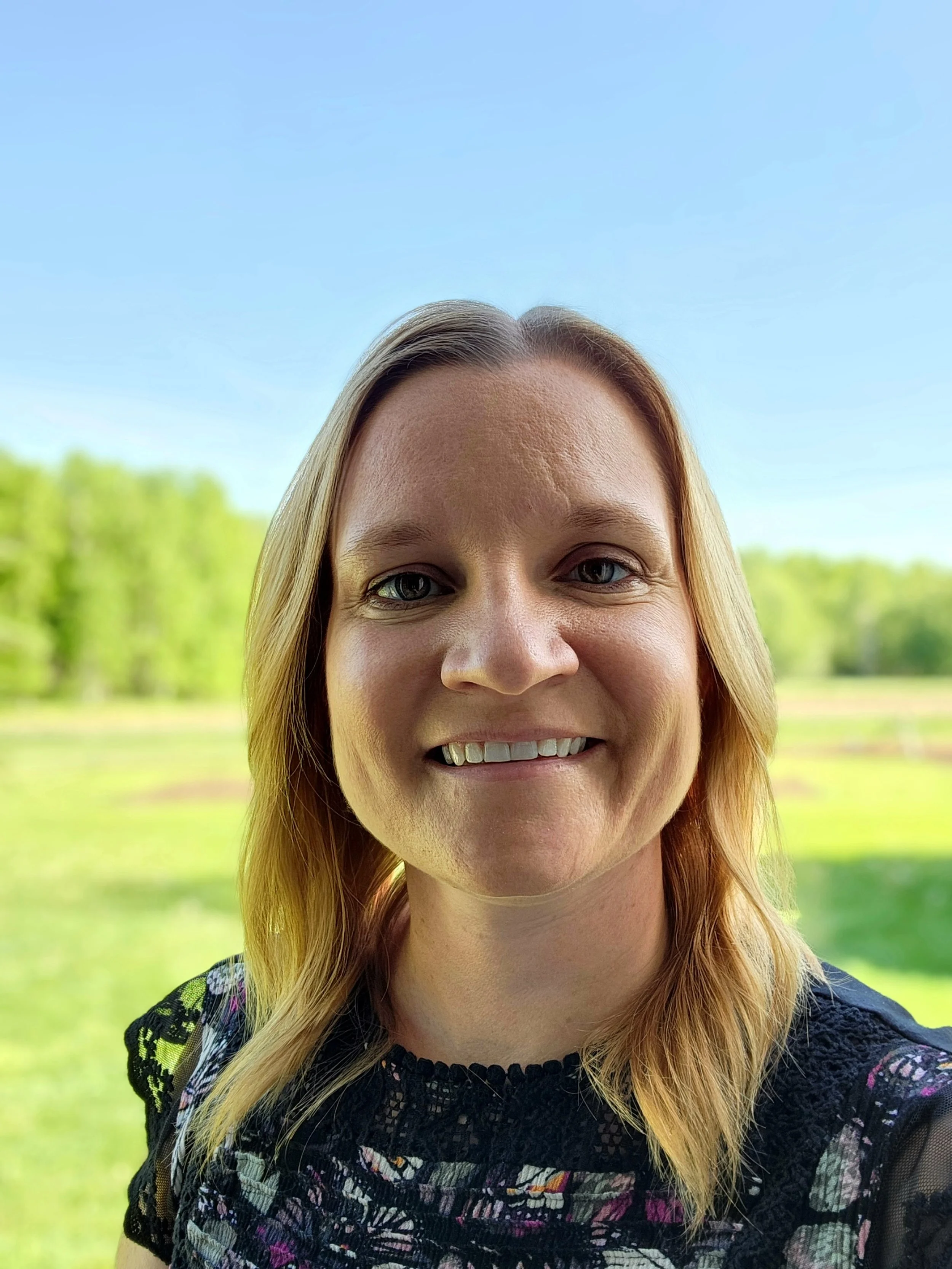 A woman smiling outdoors with a blue sky and green trees in the background.