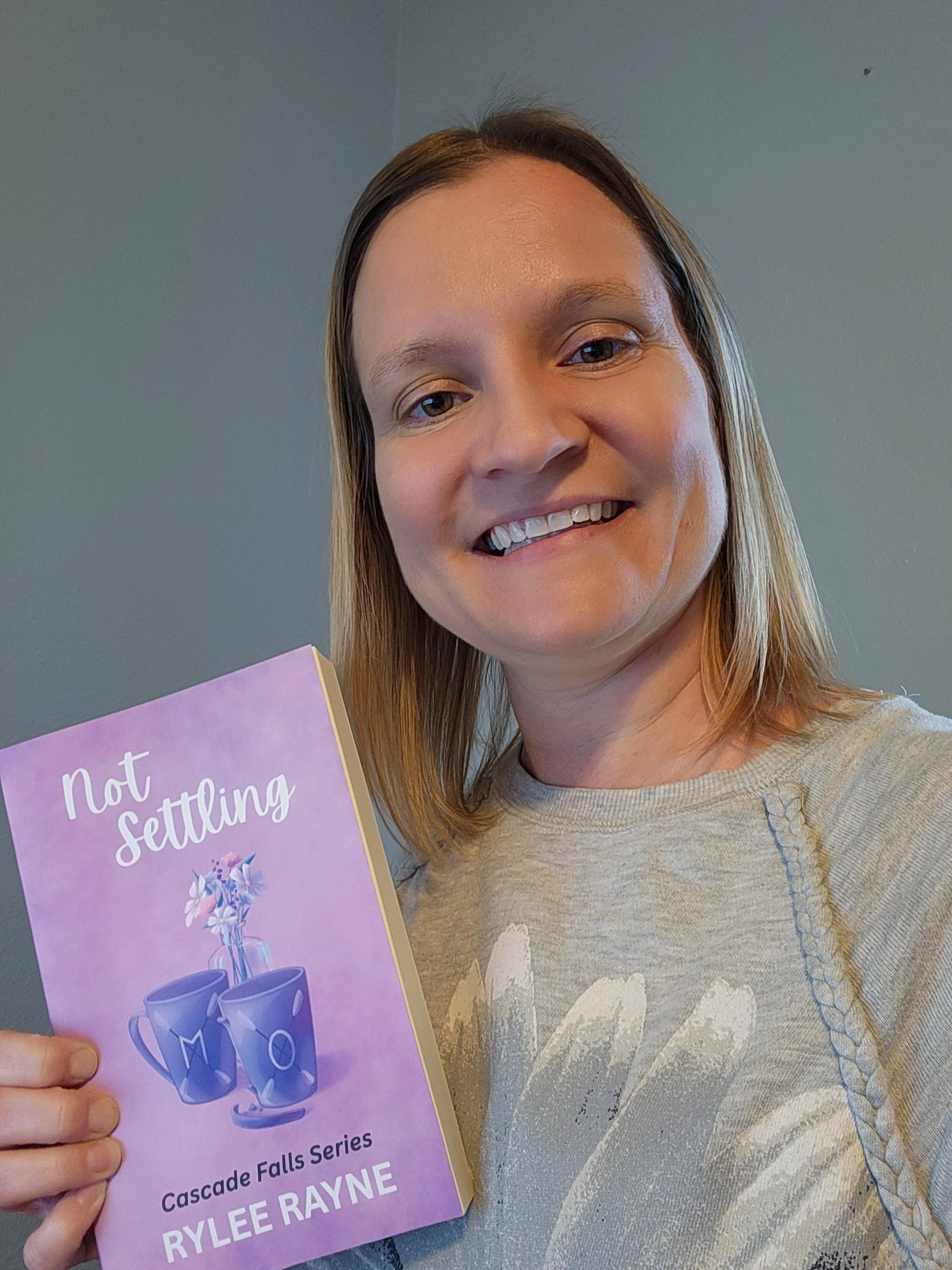 A woman with shoulder-length blonde hair smiling and holding a book titled 'Not Settling' from the Cascade Falls Series by Rylee Rayne.