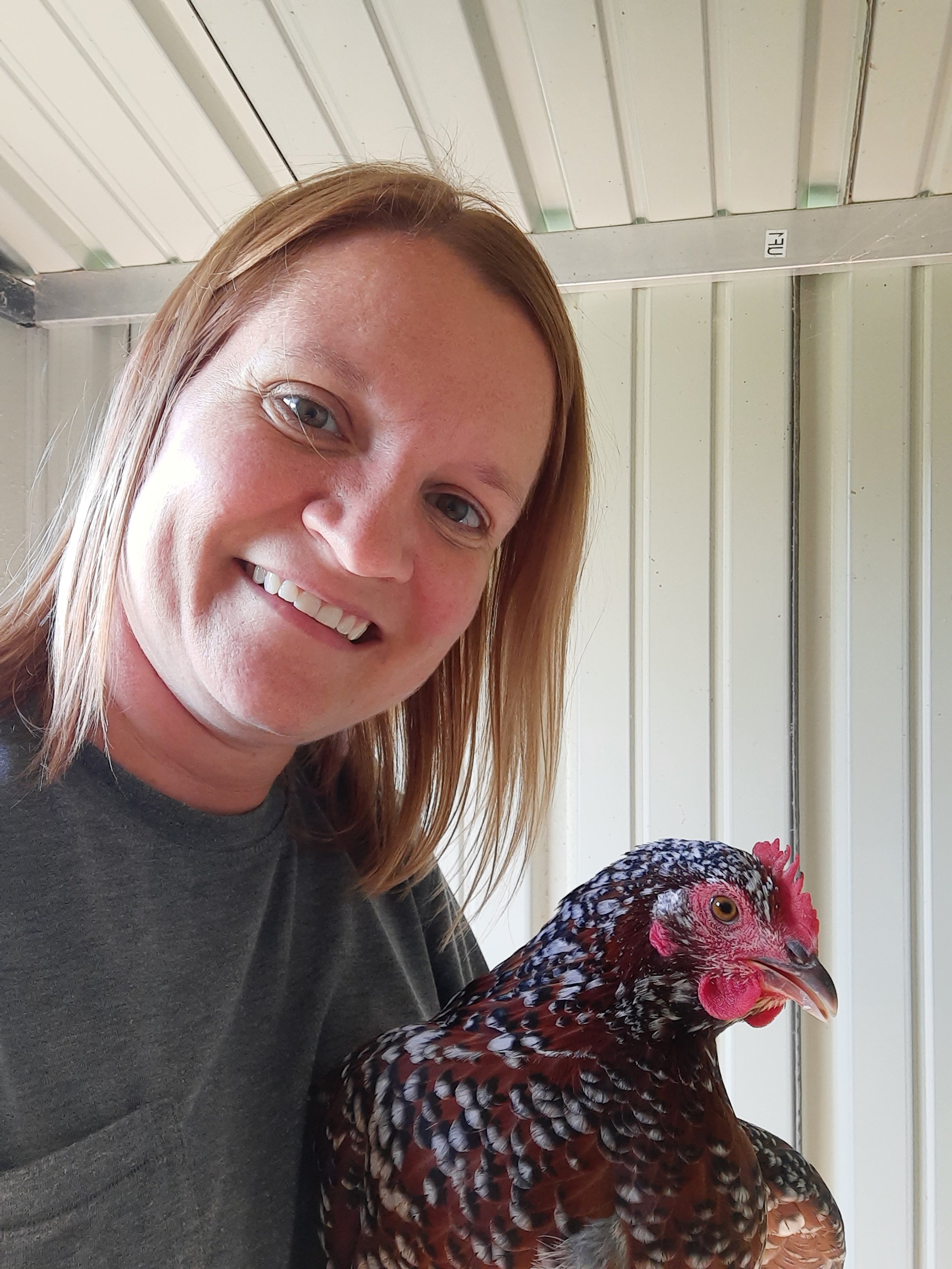 A woman with shoulder-length blonde hair smiling next to a hen inside a light-colored shed.