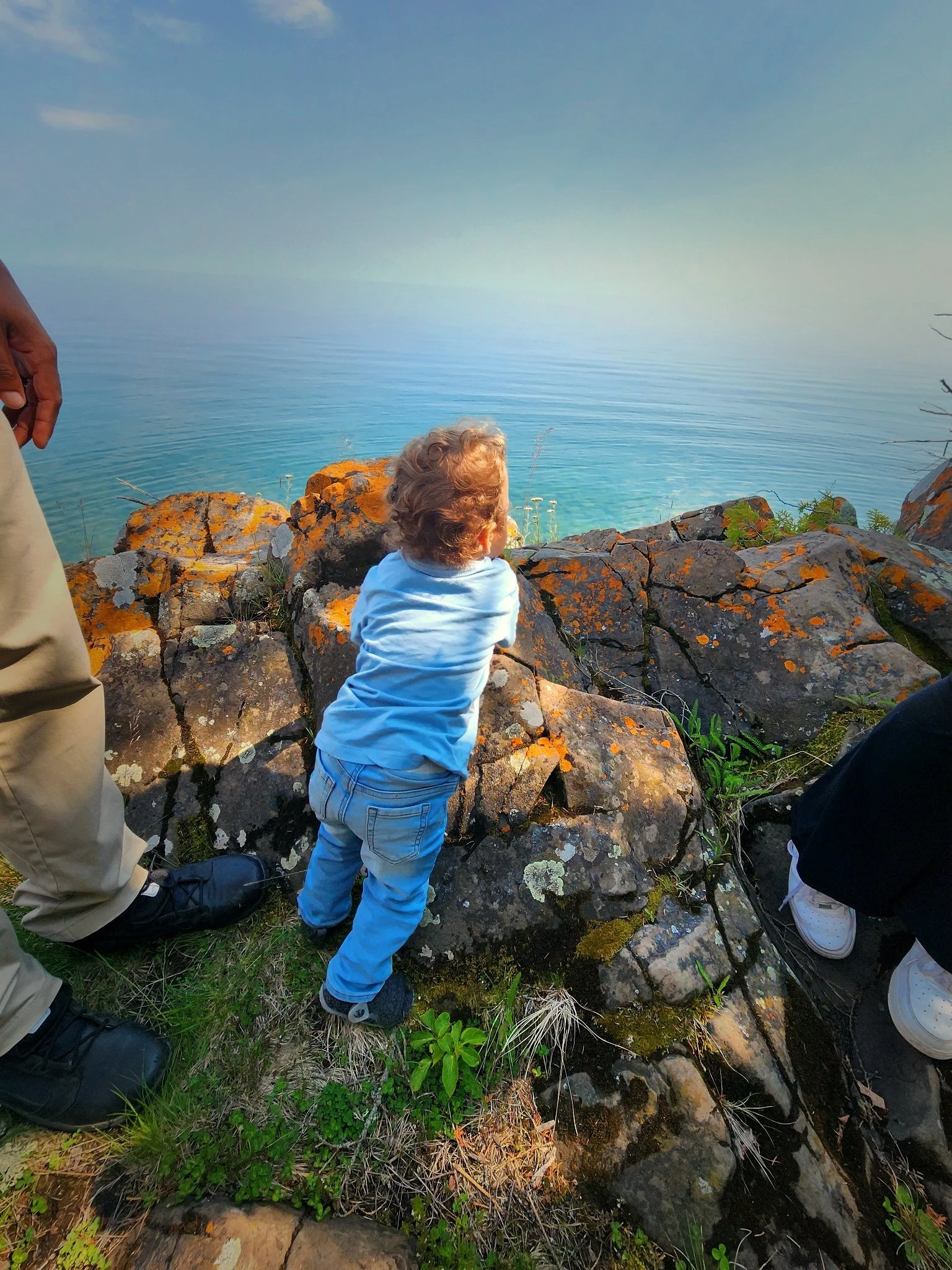 Child in a blue outfit standing on rocks, looking at a body of water, with an adult standing nearby.