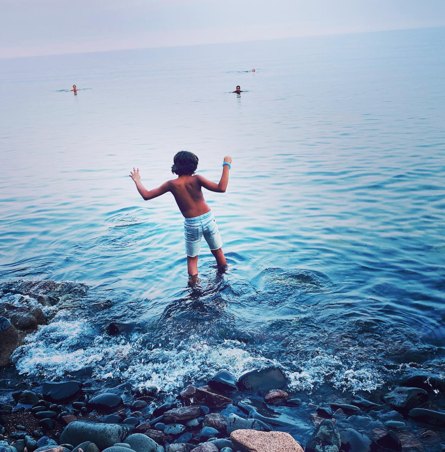 A young boy standing in shallow water at the shoreline, facing away from the camera, with his arms raised, while three people are swimming in the distance in the ocean. The shoreline has rocks and pebbles, and the water is calm.