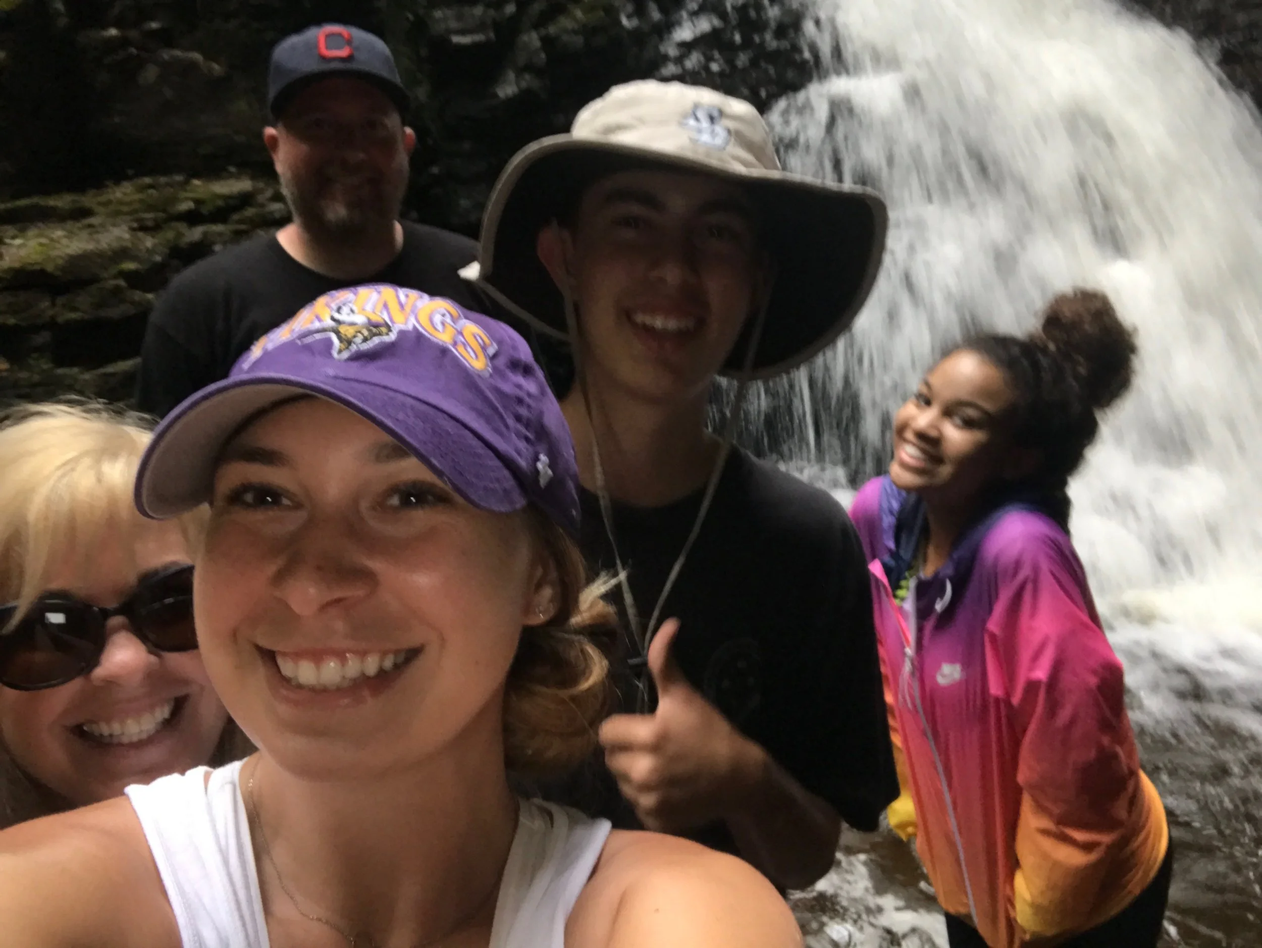 Group of five friends taking a selfie in front of a waterfall. Everyone is smiling and wearing casual outdoor clothing and hats.