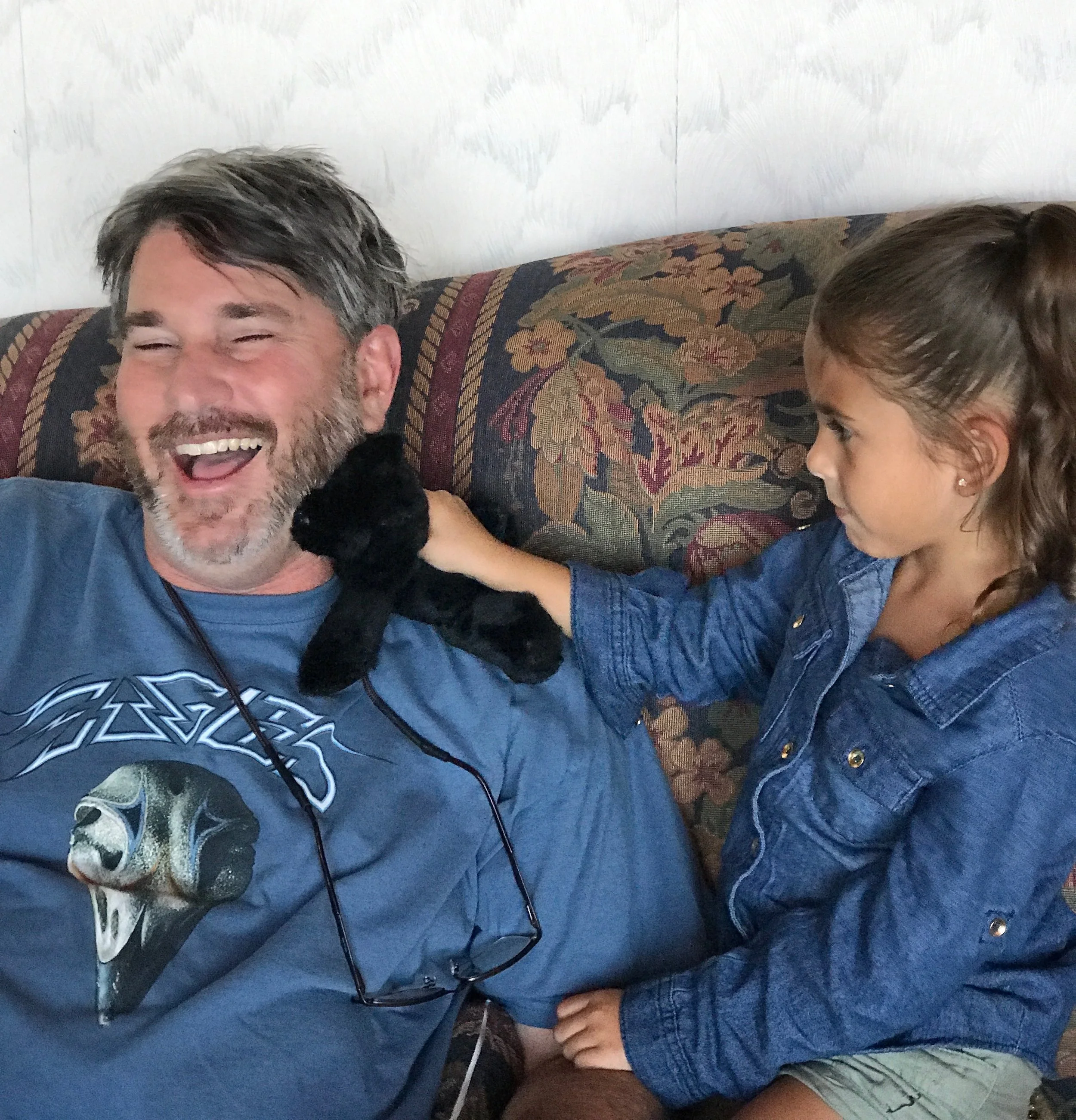 A man with gray hair and a beard laughing on a sofa, wearing a blue Metallica t-shirt and earphones, with a small black puppy touching his face; a young girl with brown hair in a ponytail and a denim jacket petting the puppy.