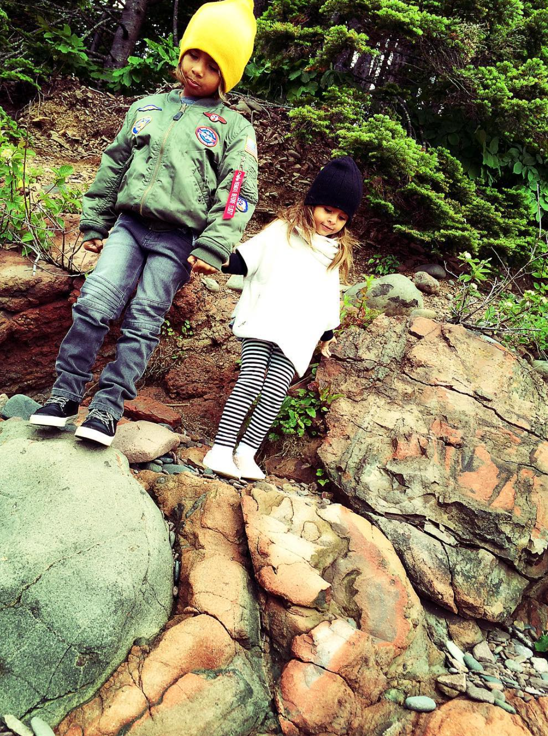 Two children, a boy and a girl, holding hands on a rocky trail outdoors surrounded by greenery, with trees and bushes in the background, during daytime.