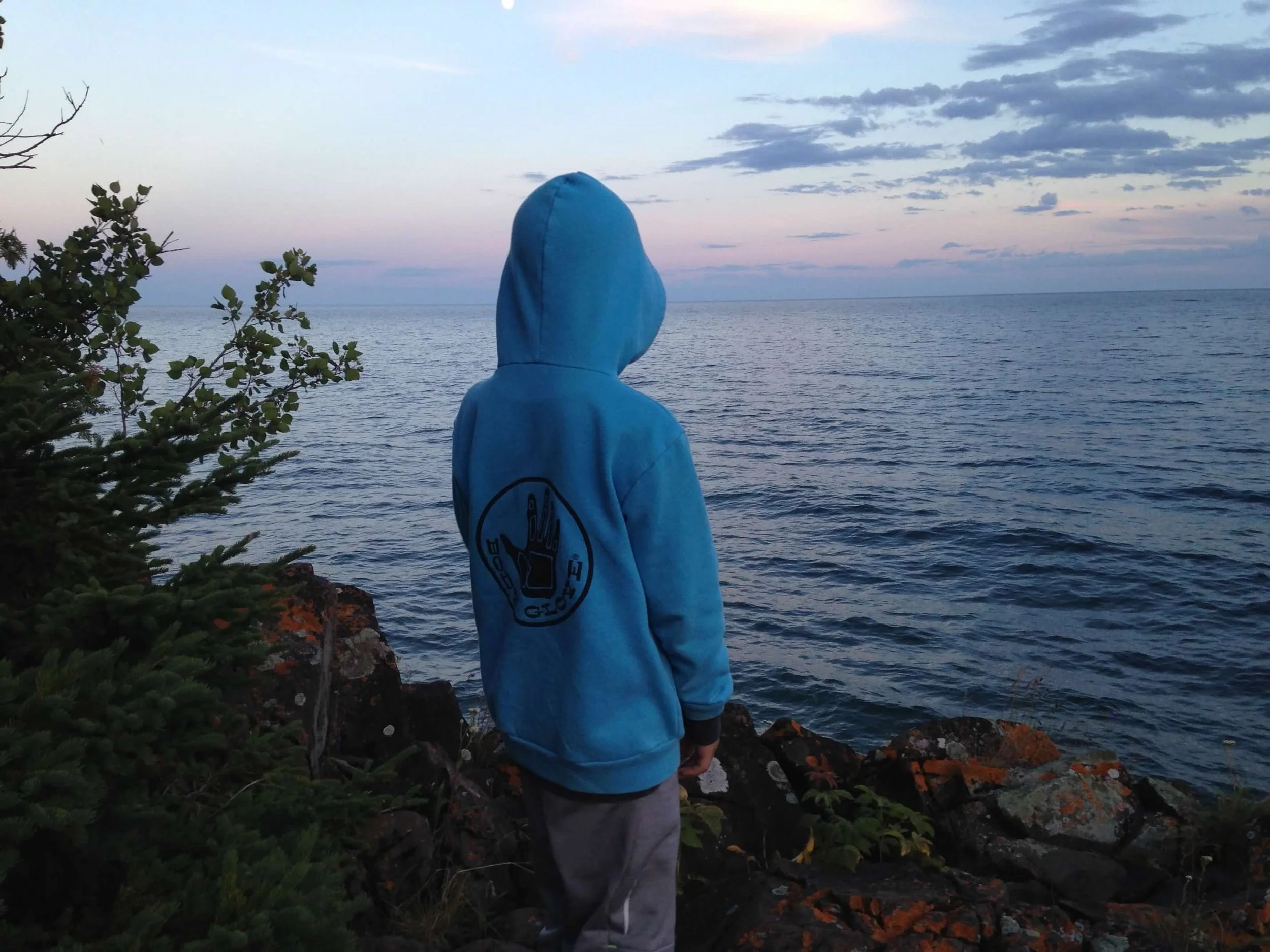 A child wearing a blue hoodie with a graphic on the back, standing on rocky shoreline overlooking the ocean at dusk.