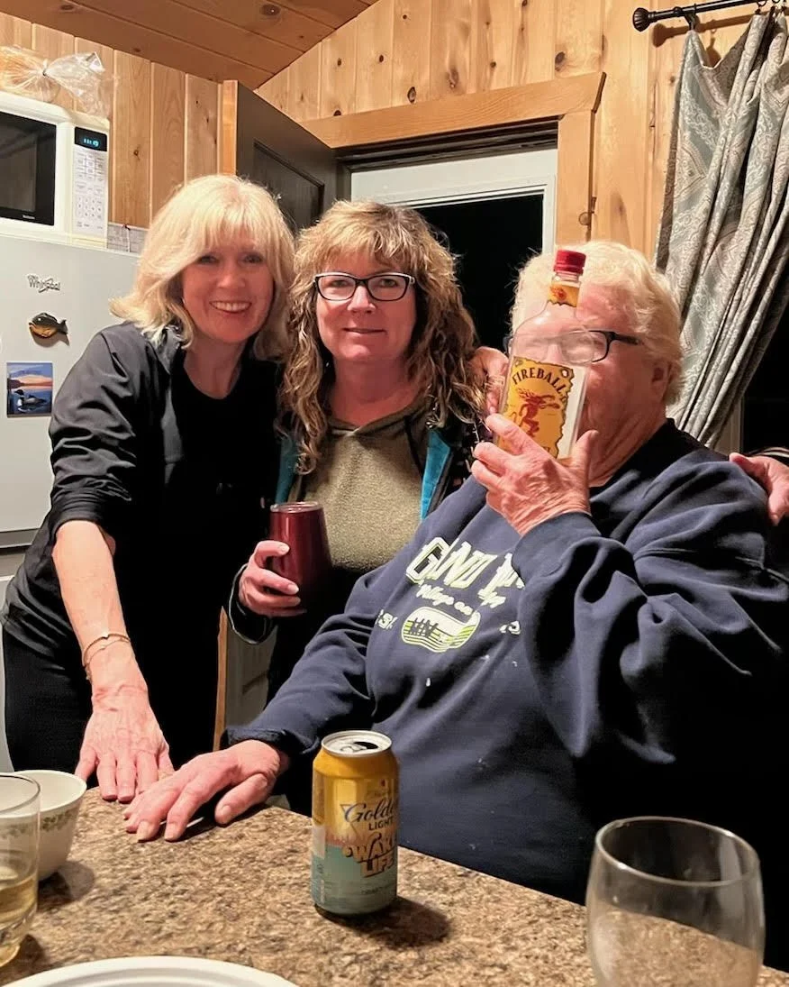 Three women gathered at a kitchen counter, smiling and holding drinks, with a man sitting at the table holding a bottle of Fireball whiskey.