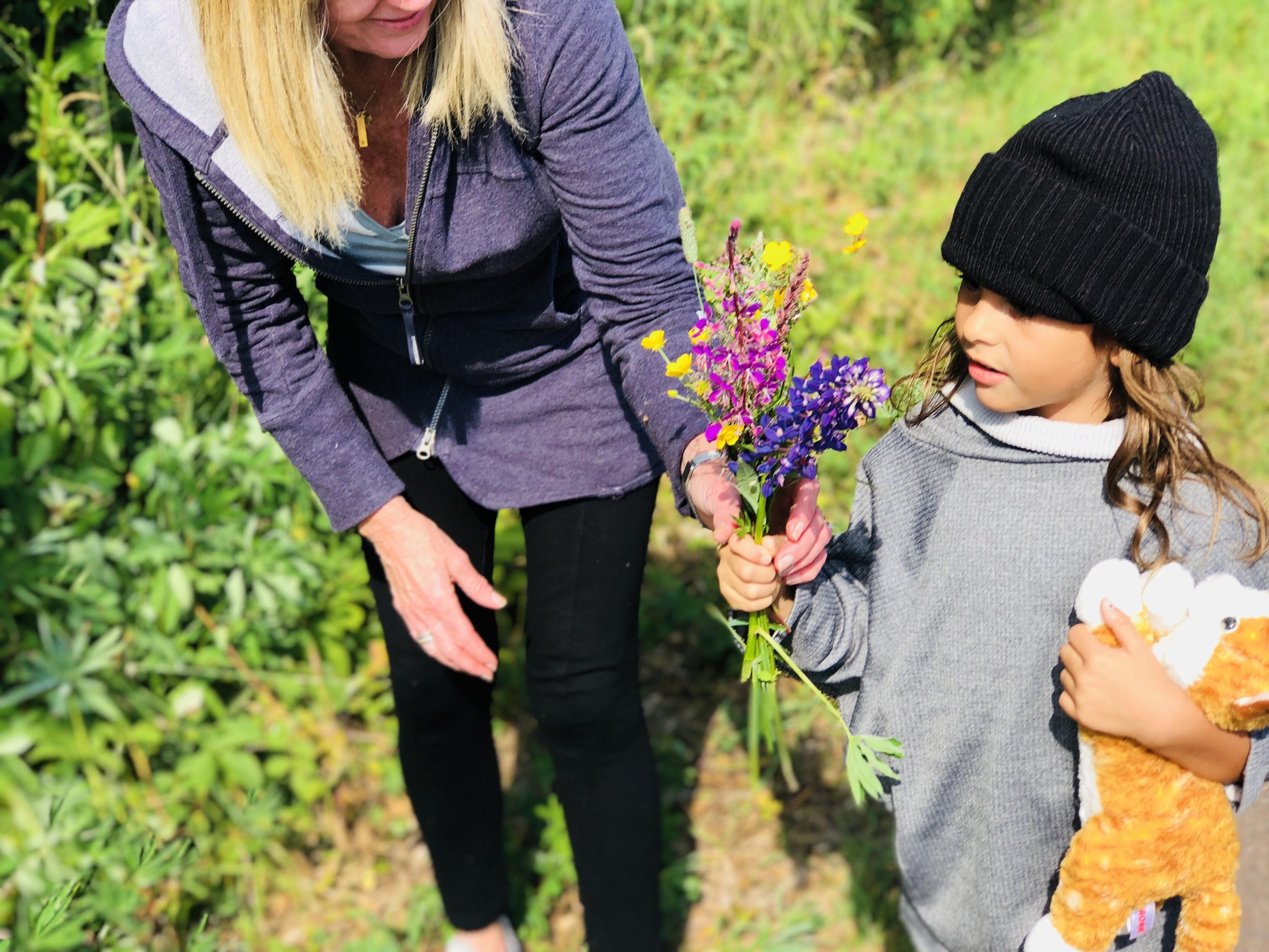 A woman giving flowers to a young girl outdoors in a green garden, with the girl holding a stuffed animal.