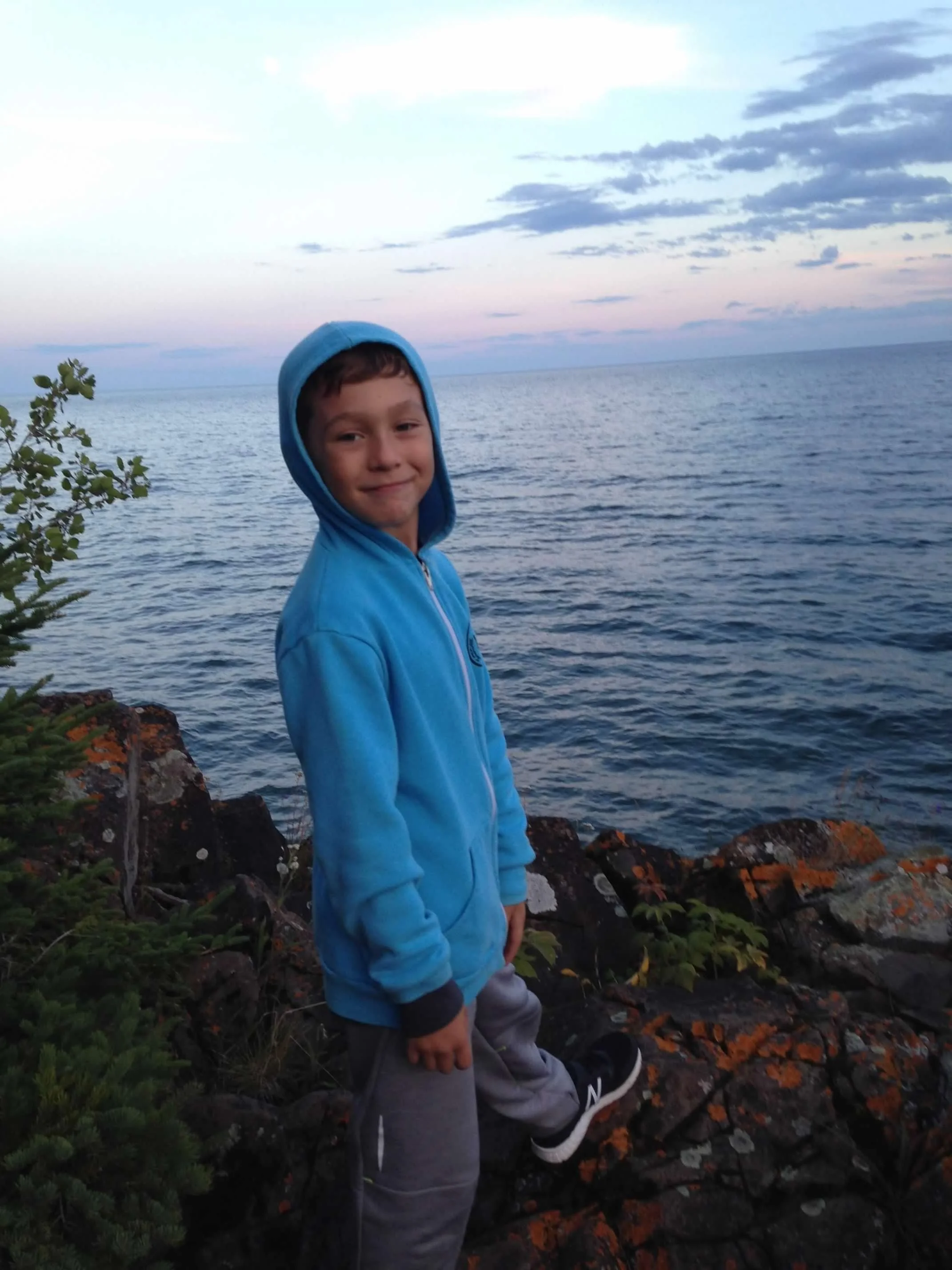 Young boy wearing a blue hoodie and gray pants standing on rocky shoreline by the water during sunset.