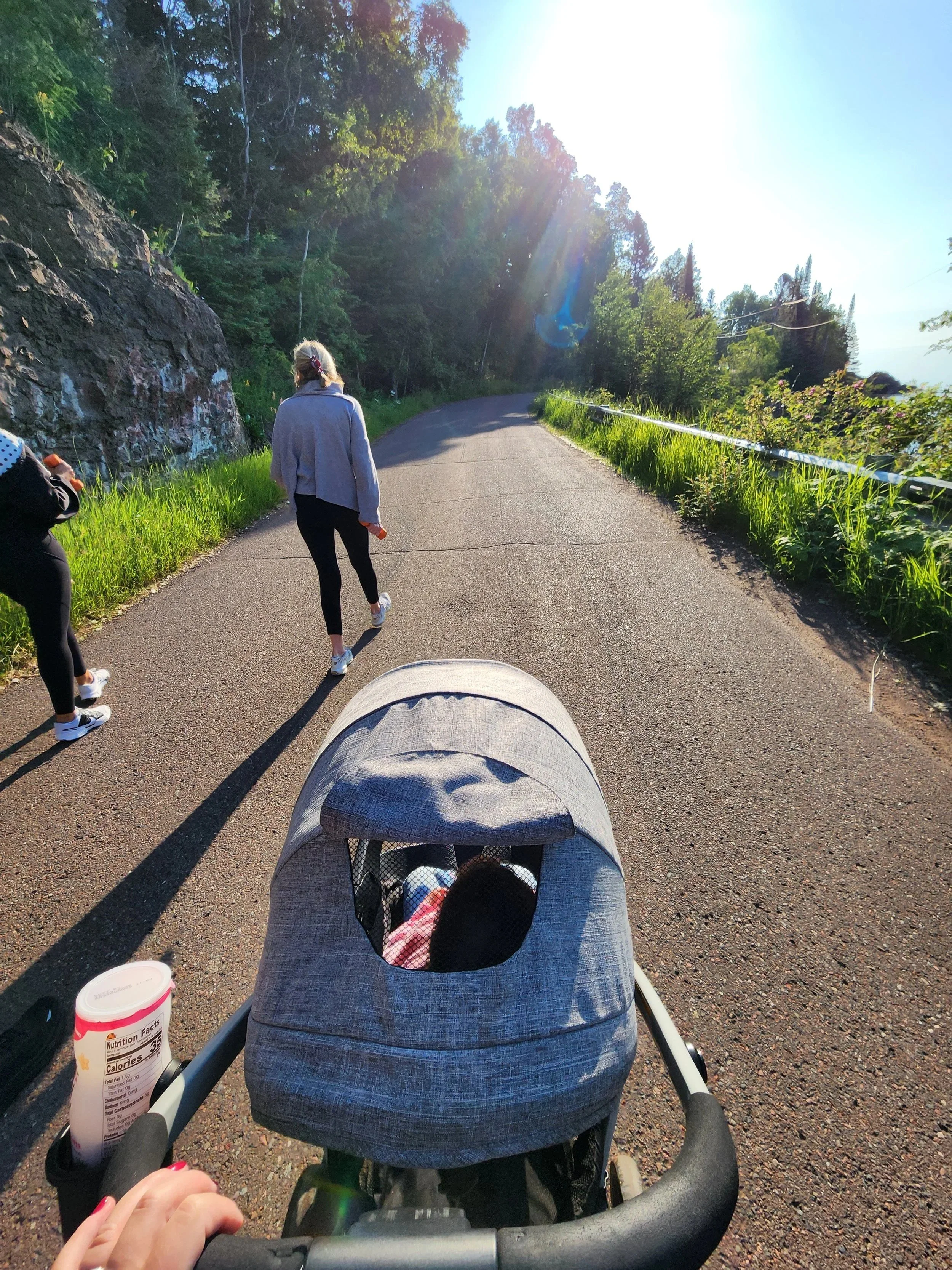 View from a stroller on a paved road with two women walking ahead, surrounded by lush green trees and grass on a sunny day.