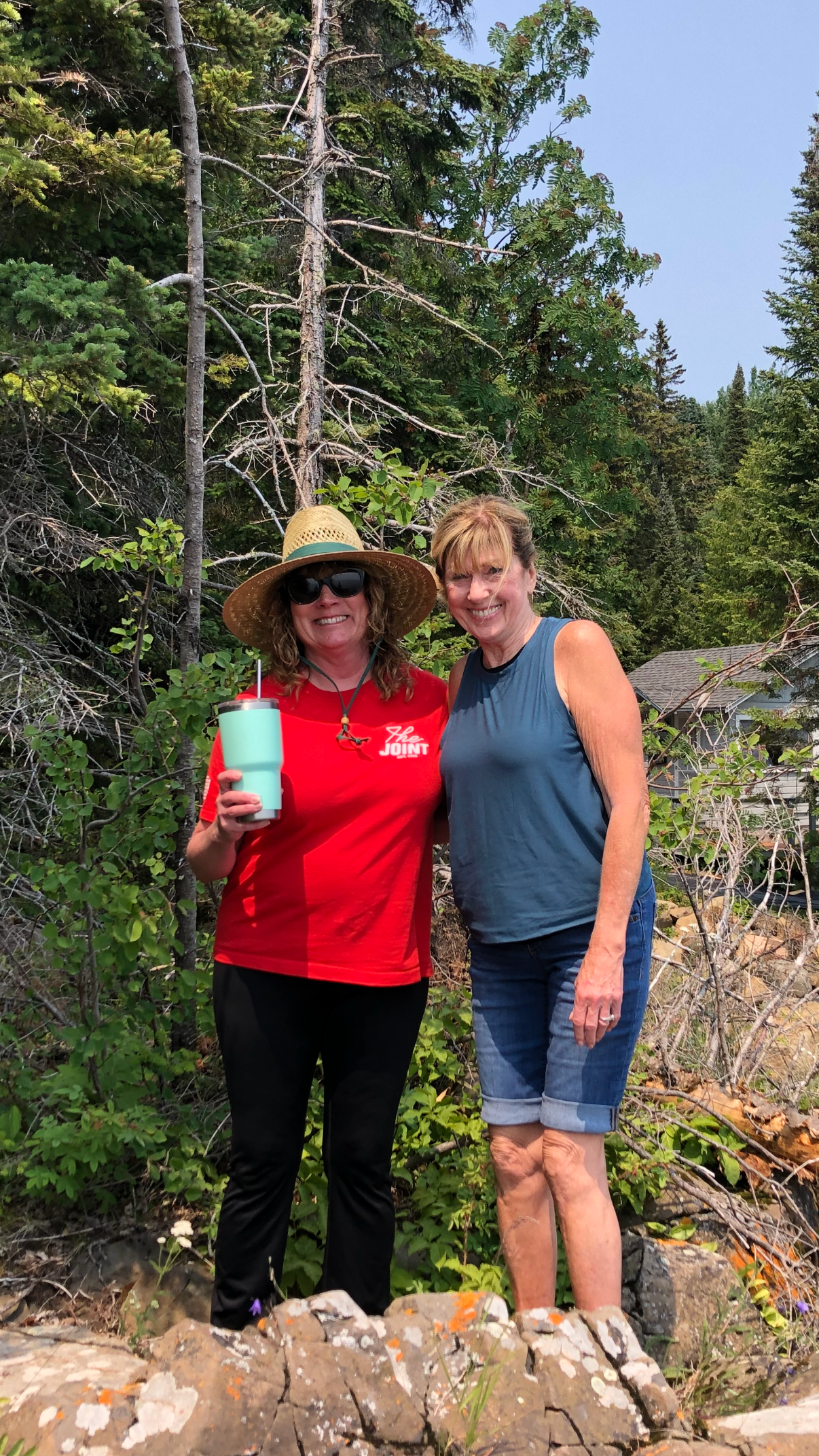 Two women smiling outdoors in front of a wooded area, one wearing a wide-brimmed hat, sunglasses, and a red shirt, holding a tumbler, and the other in a sleeveless blue top and denim shorts.
