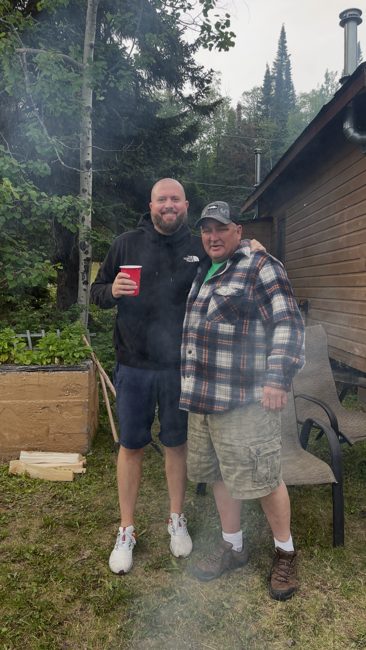 Two men standing outdoors in a backyard, smiling, one holding a red cup, with trees and a wooden house in the background.