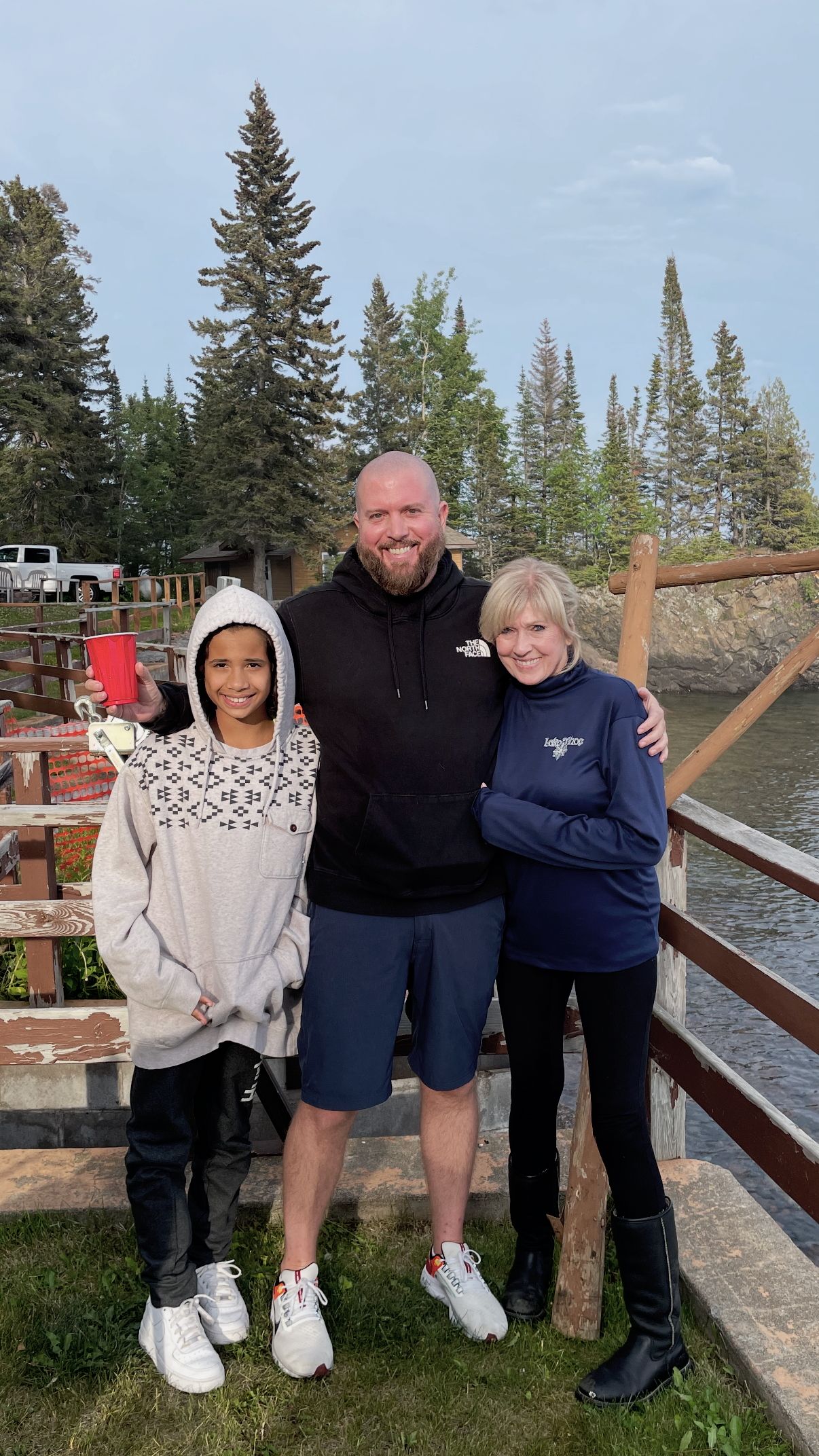 Three people smiling outdoors near water and trees, holding a red cup. Woman on right wearing a blue jacket, man in sunglasses wearing a black hoodie, girl on left in a hoodie with a pattern.