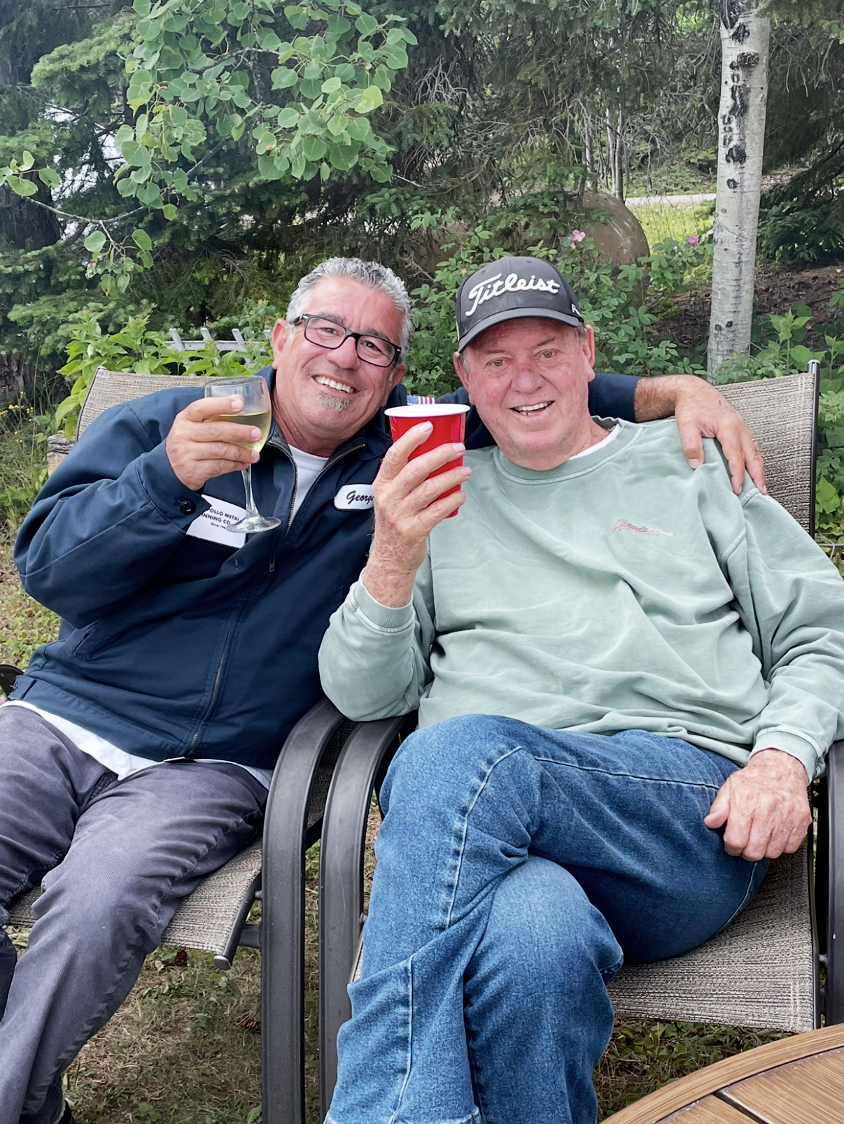 Two men smiling and enjoying drinks outside, one with a glass of wine and the other with a red cup, sitting on outdoor chairs surrounded by trees and greenery.