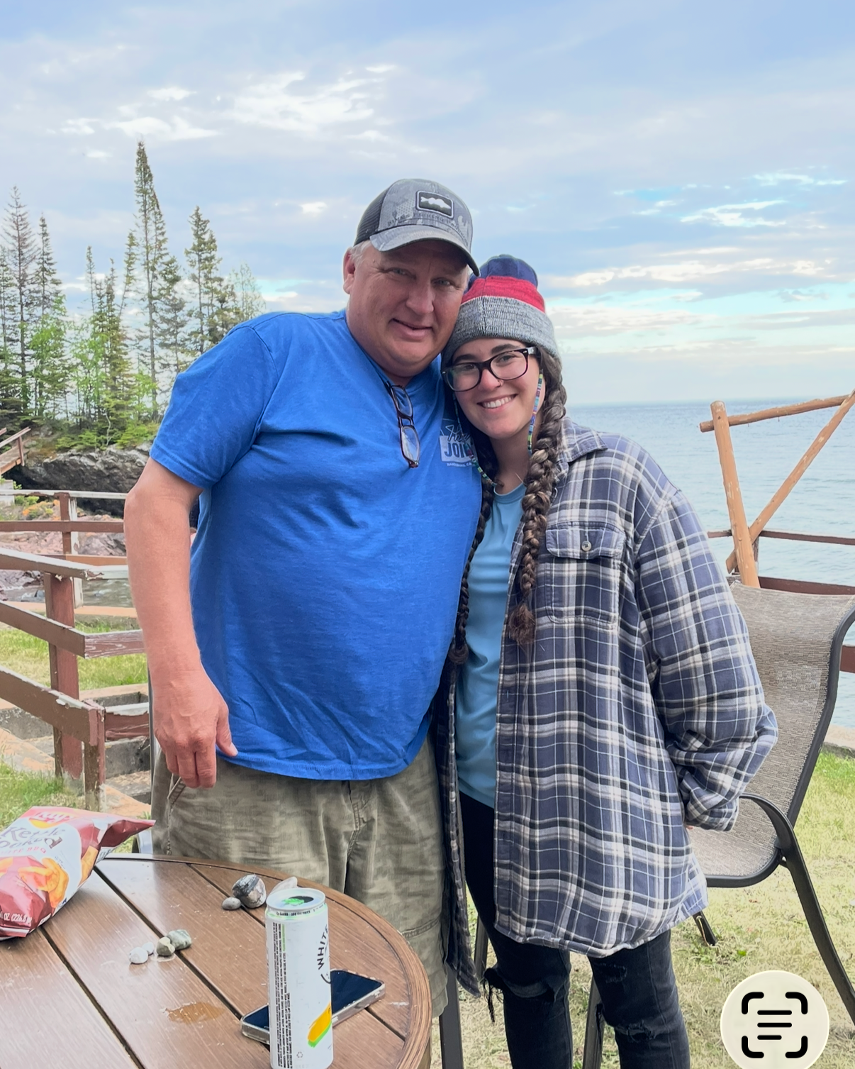 A man and woman are standing outdoors near a body of water with trees in the background. The man is wearing a blue shirt and a gray cap, and the woman is wearing a gray beanie, glasses, and a plaid jacket. They are smiling and hugging each other.