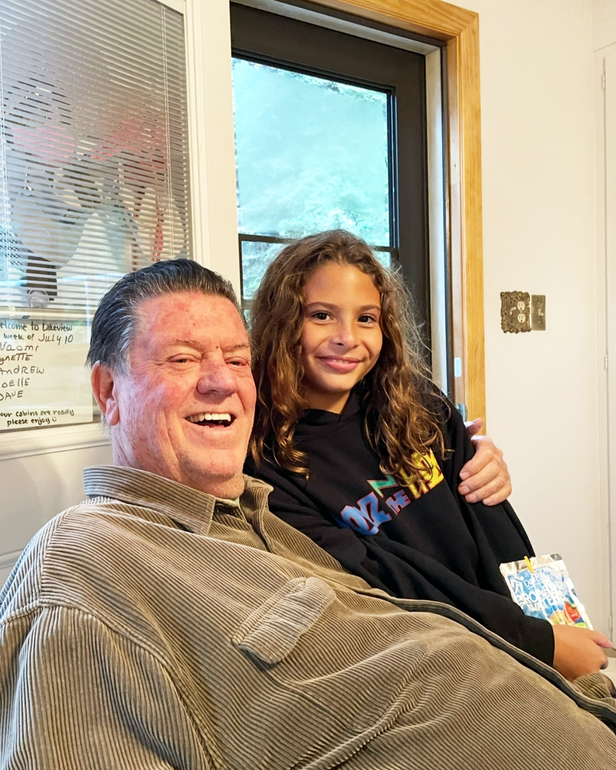 A man and a girl sitting close together, smiling at the camera indoors near a window with a wooden frame. The man has short dark hair and is wearing a tan corduroy shirt. The girl has curly light brown hair and is wearing a black hoodie with colorful