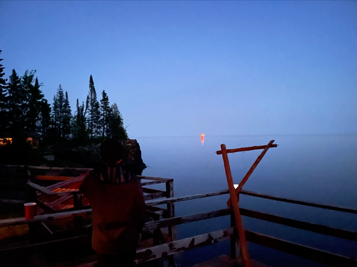 Person standing on a wooden deck looking out over a calm body of water during dusk with a lighthouse in the distance and a small forested island to the left.
