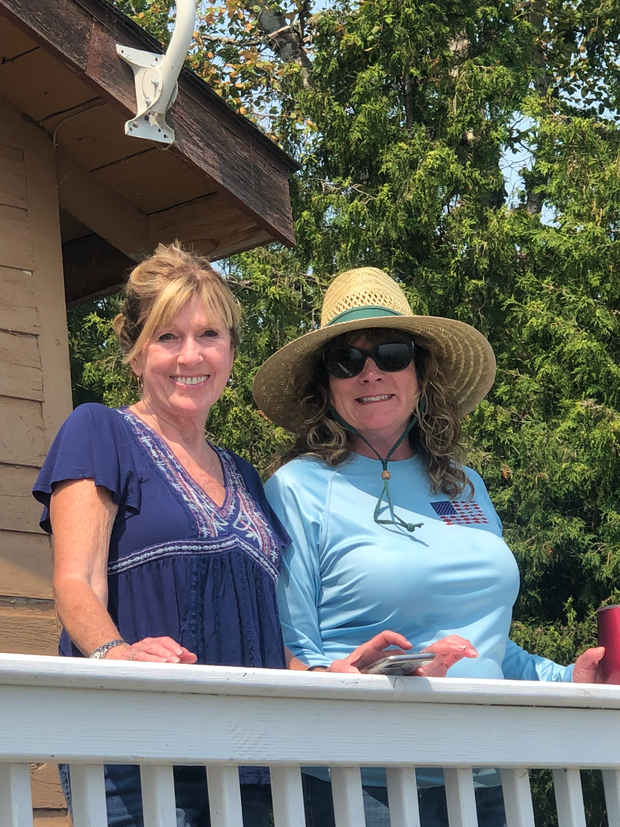 Two women smiling on a porch. One is wearing a blue top, and the other is wearing a light blue shirt, large wide-brimmed hat, and sunglasses. They are outside near a tree and a building with beige siding.