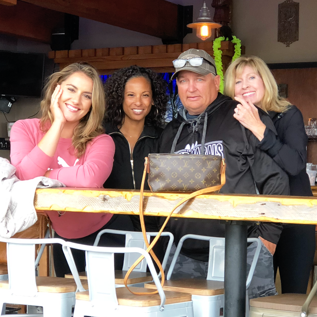 Four people, three women and one man, are gathered around a table in a cozy restaurant. They are smiling and looking at the camera, with some leaning on their hands. The woman on the left has long blonde hair and is wearing a pink sweatshirt. The wom