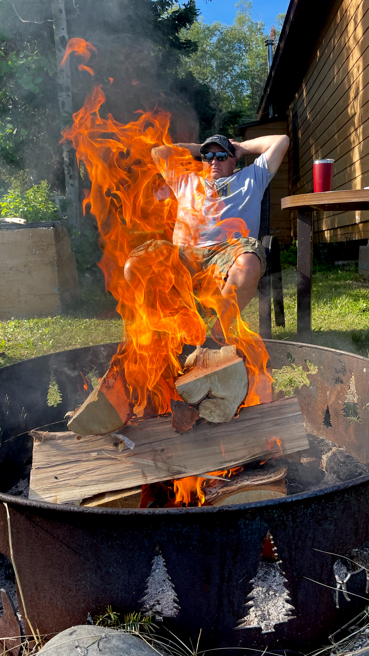 Man relaxing outdoors in a chair next to a fire pit with burning logs.