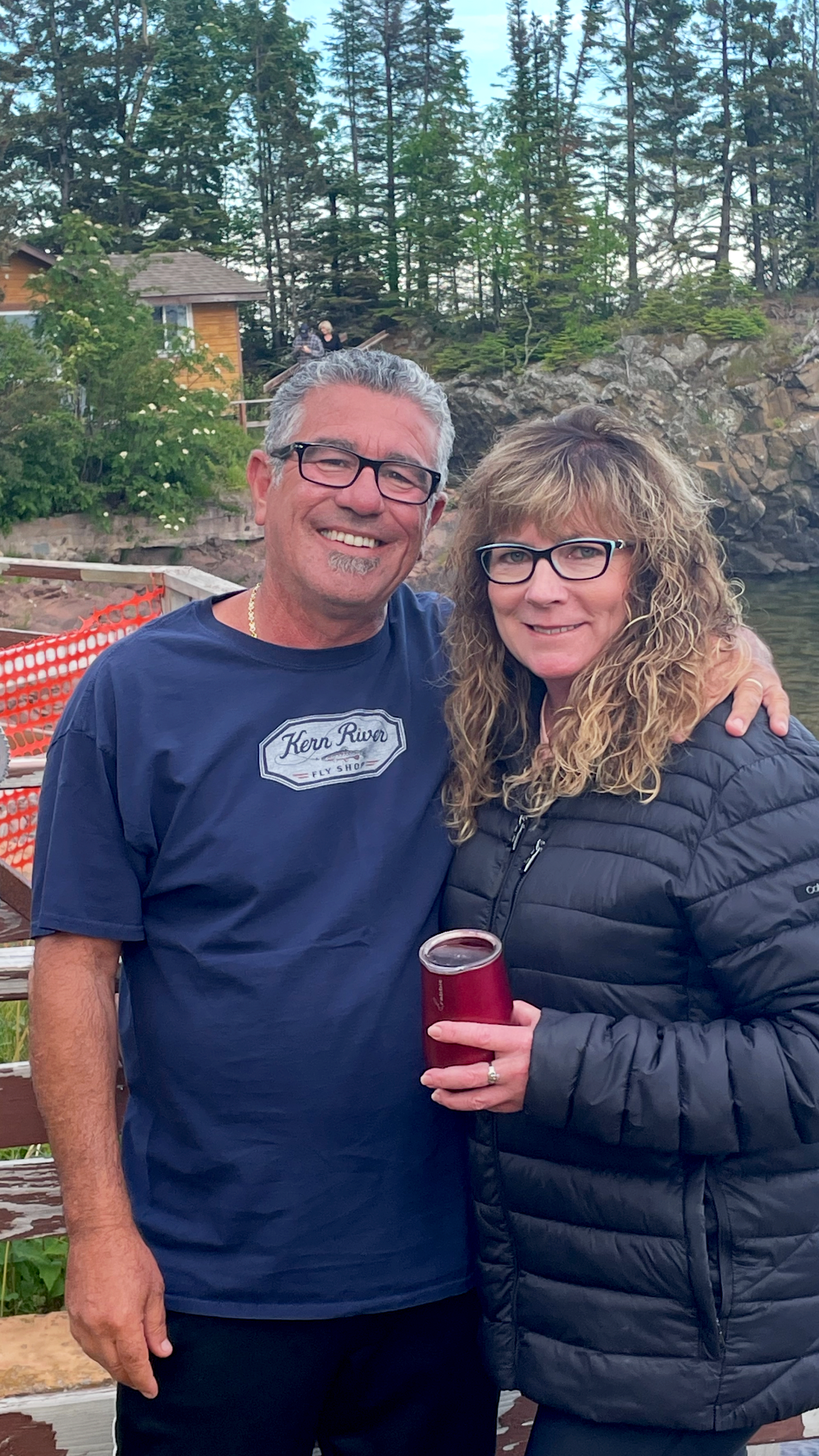 A smiling middle-aged man and woman standing outdoors near a body of water, with trees and a house in the background. The man is wearing glasses and a blue T-shirt. The woman is wearing glasses and a black jacket, holding a red can.