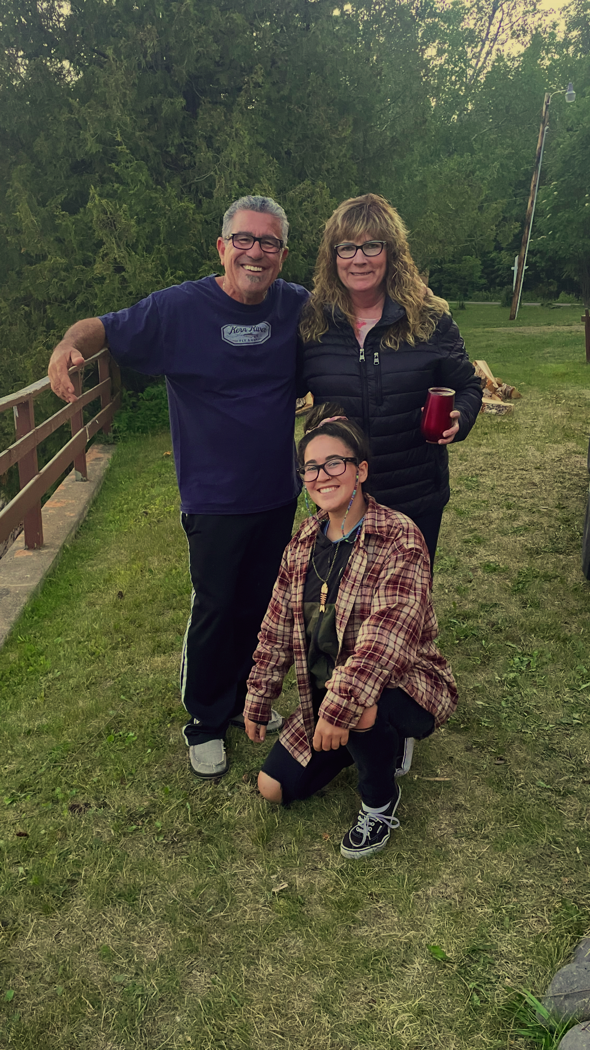 Three people smiling outdoors on a grassy area with trees in the background, two adults standing and one kneeling, all wearing casual clothing and glasses.