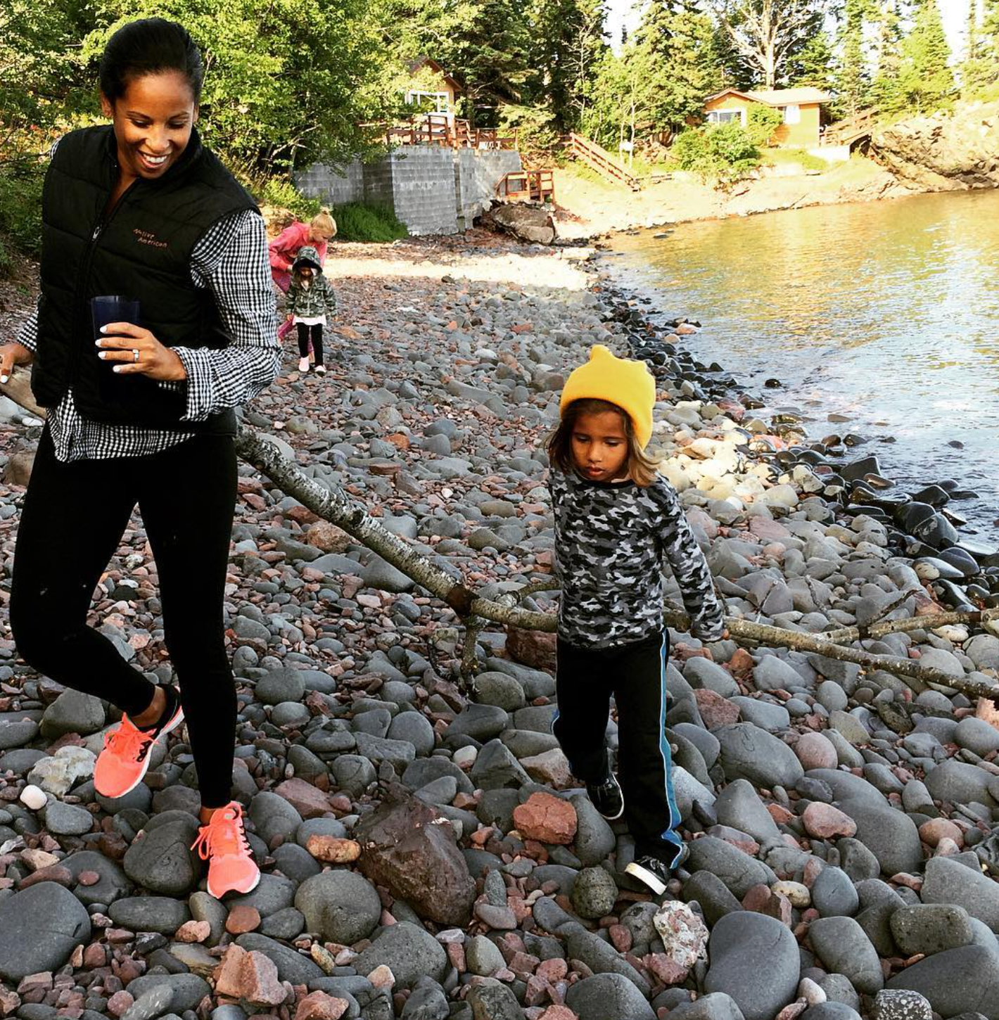 Woman and young girl walking on a rocky beach near a body of water. The woman is smiling and holding a cup, while the girl, wearing a yellow hat, looks down at the rocks.