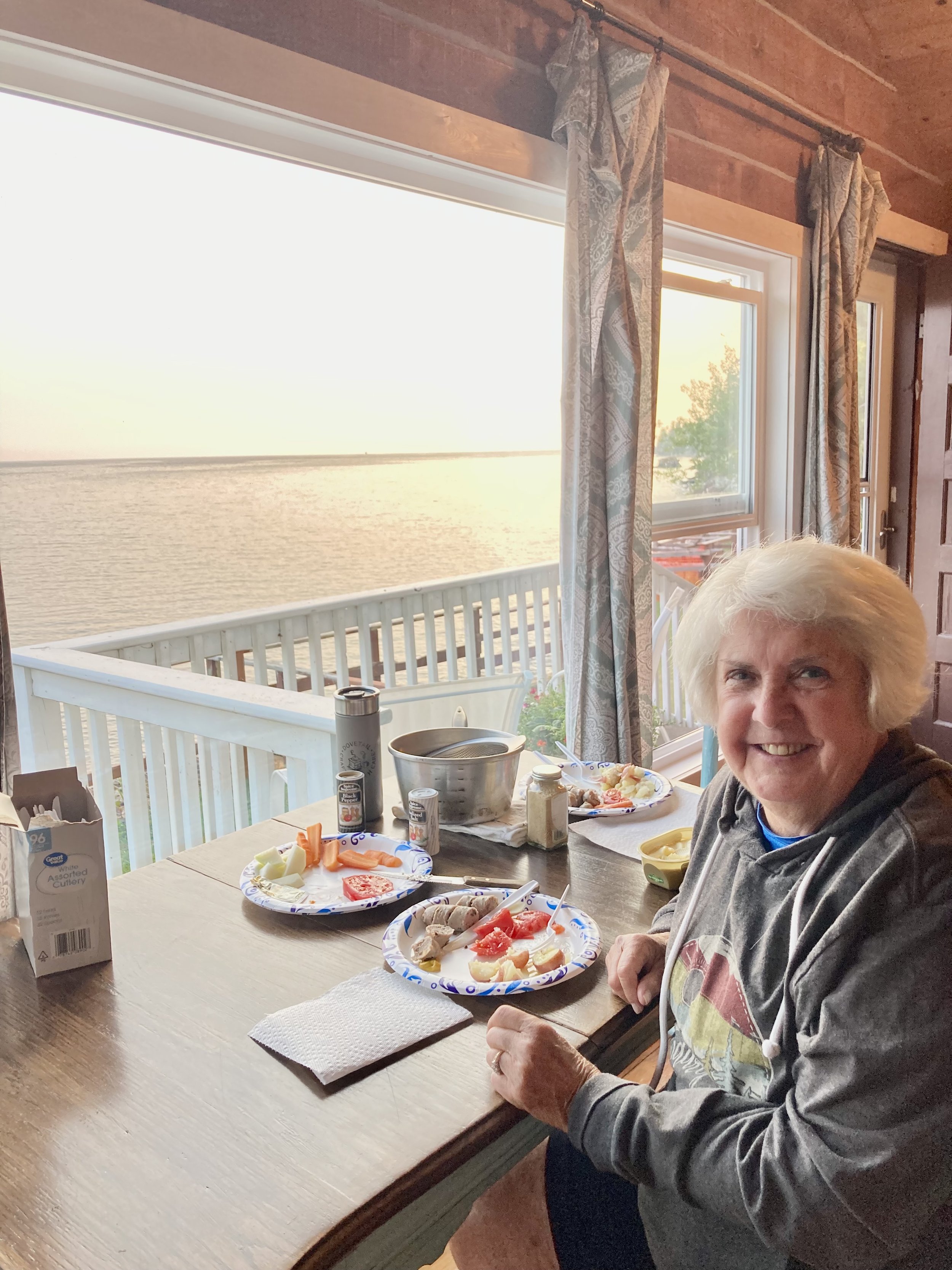 A smiling elderly woman sitting at a dining table by a large window overlooking a body of water at sunset. The table has paper plates with partially eaten food, condiments, and a napkin.